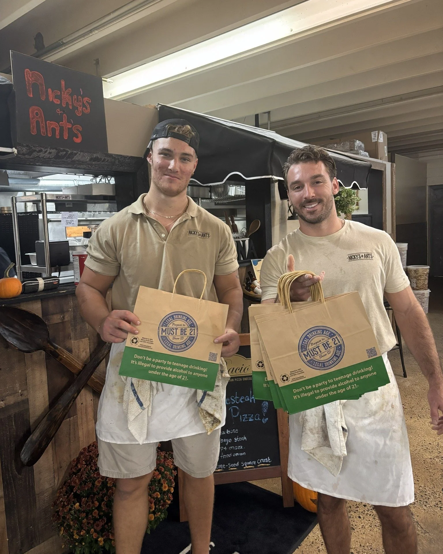 Two young men wearing beige shirts holding paper bags with a message about underage drinking in a store or cafe setting.