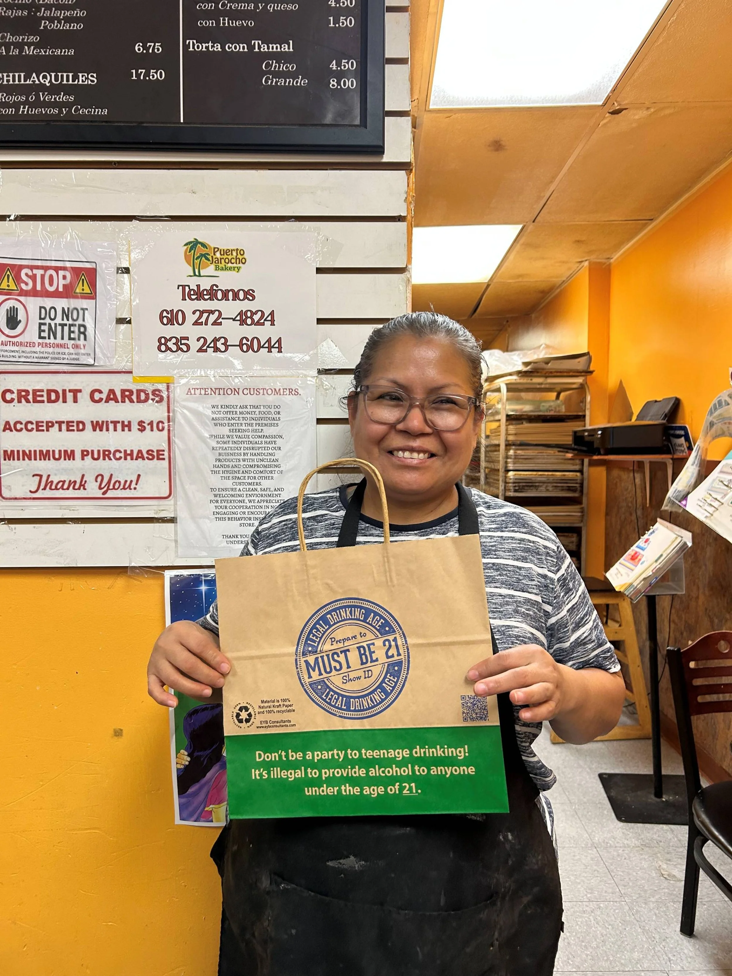 Smiling woman in gray striped shirt and glasses holding a paper bag with a legal drinking age sign inside. The background features orange and white walls, a menu board, and various signs including a "Stop Do Not Enter" sign and a notice about credit 