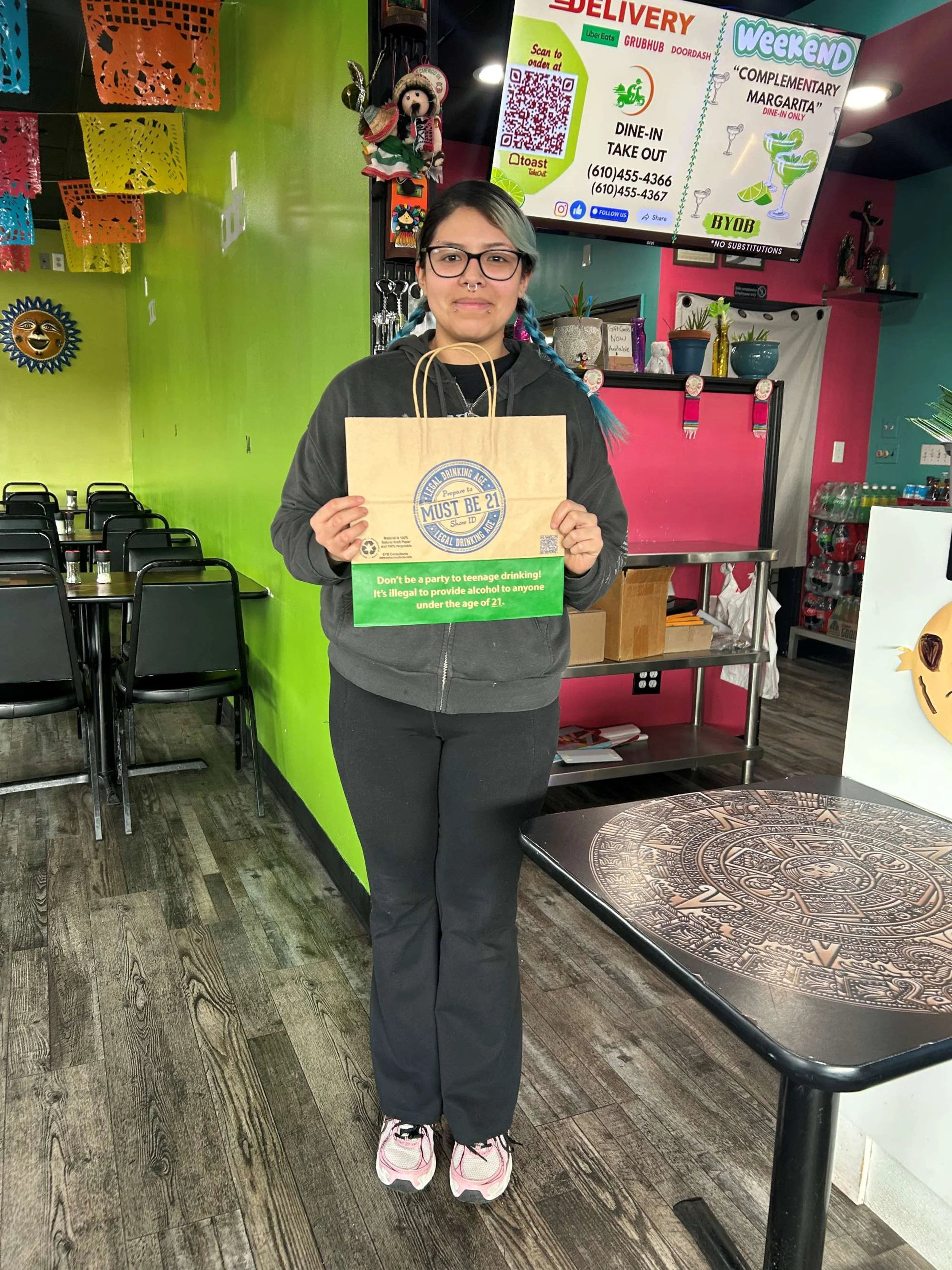 A young woman with glasses and multicolored braided hair stands inside a colorful restaurant, holding a paper bag. The restaurant's decor includes bright green and pink walls, decorative Papel Picado banners, and various ornaments. A large screen beh