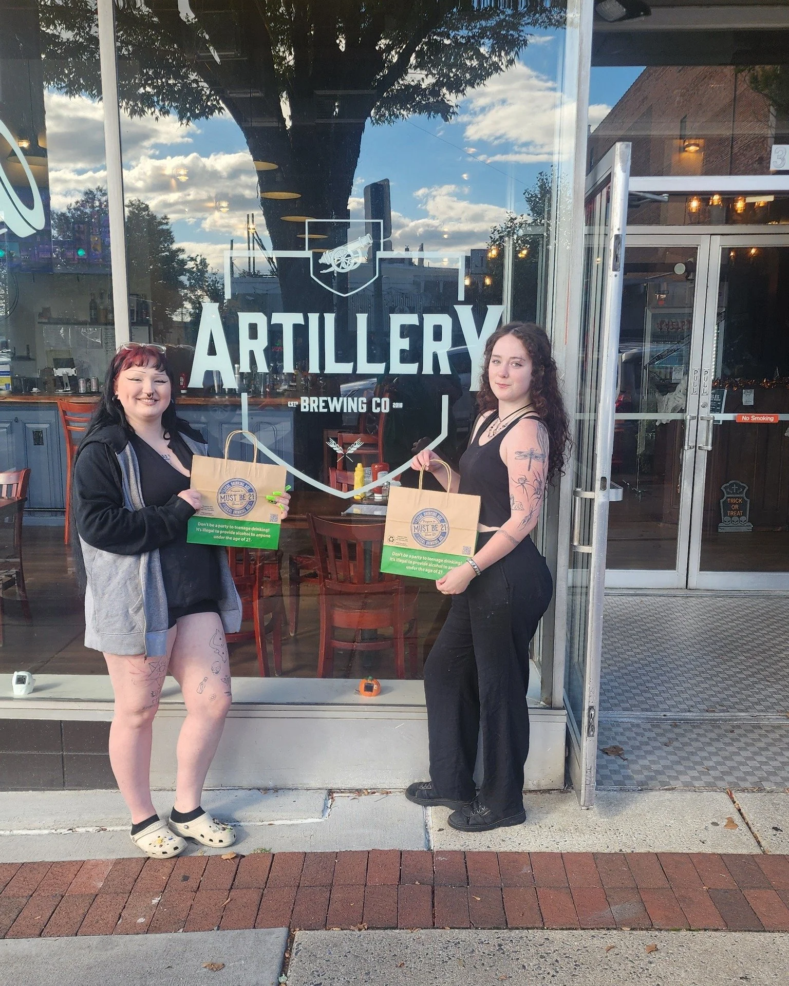 Two women standing outside the entrance of a brewery called Artillery Brewing Co., holding paper bags. The woman on the left has black hair with red highlights, tattoos on her legs, and is wearing a black top, gray hoodie, and Crocs. The woman on the