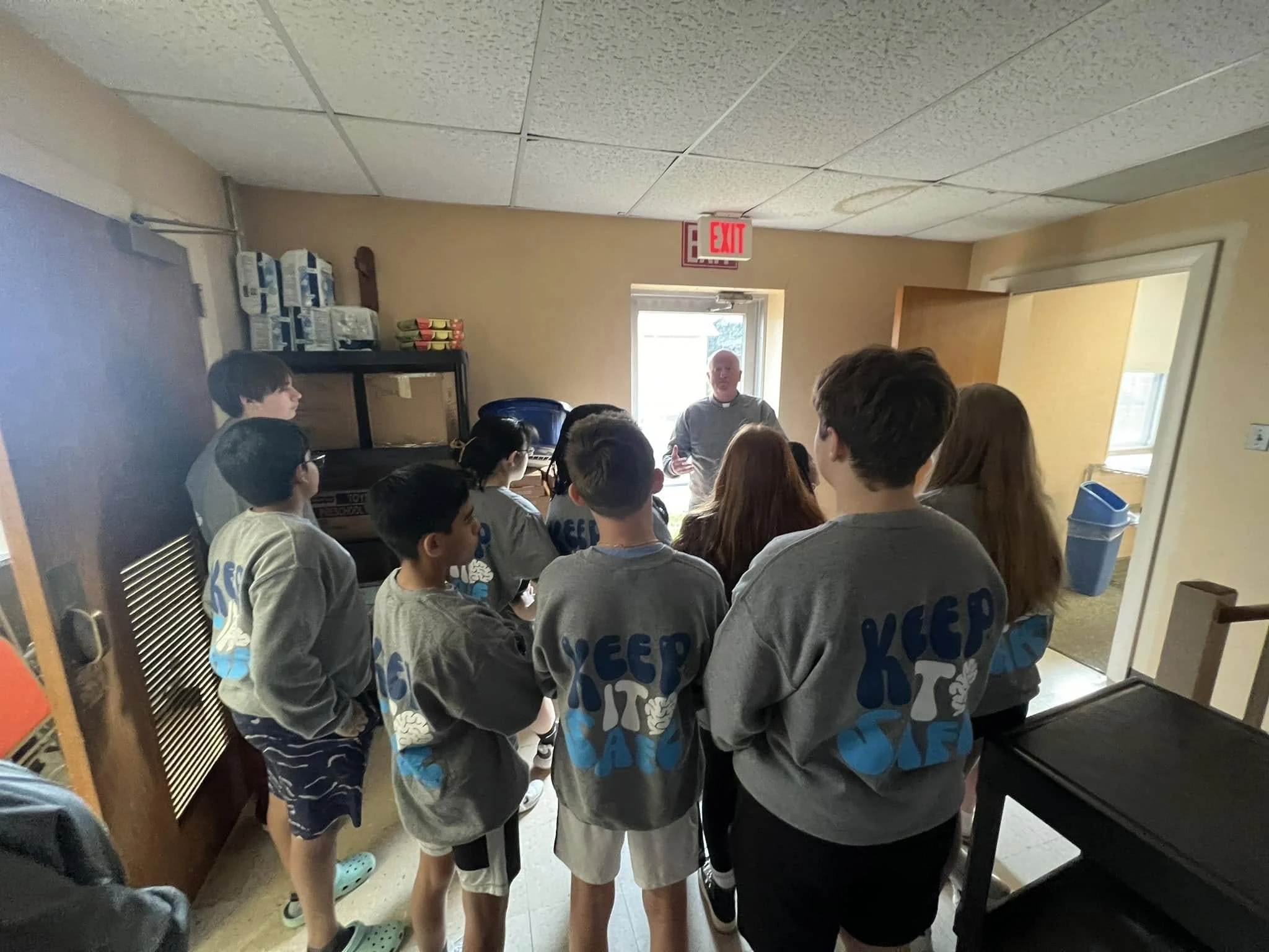 A group of children wearing gray sweatshirts with blue text gathering around a man in a room with a small door leading outside.