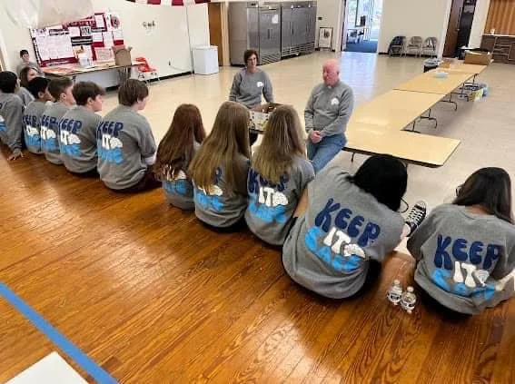 Group of students sitting on the floor in a classroom, facing two adults who are talking to them.