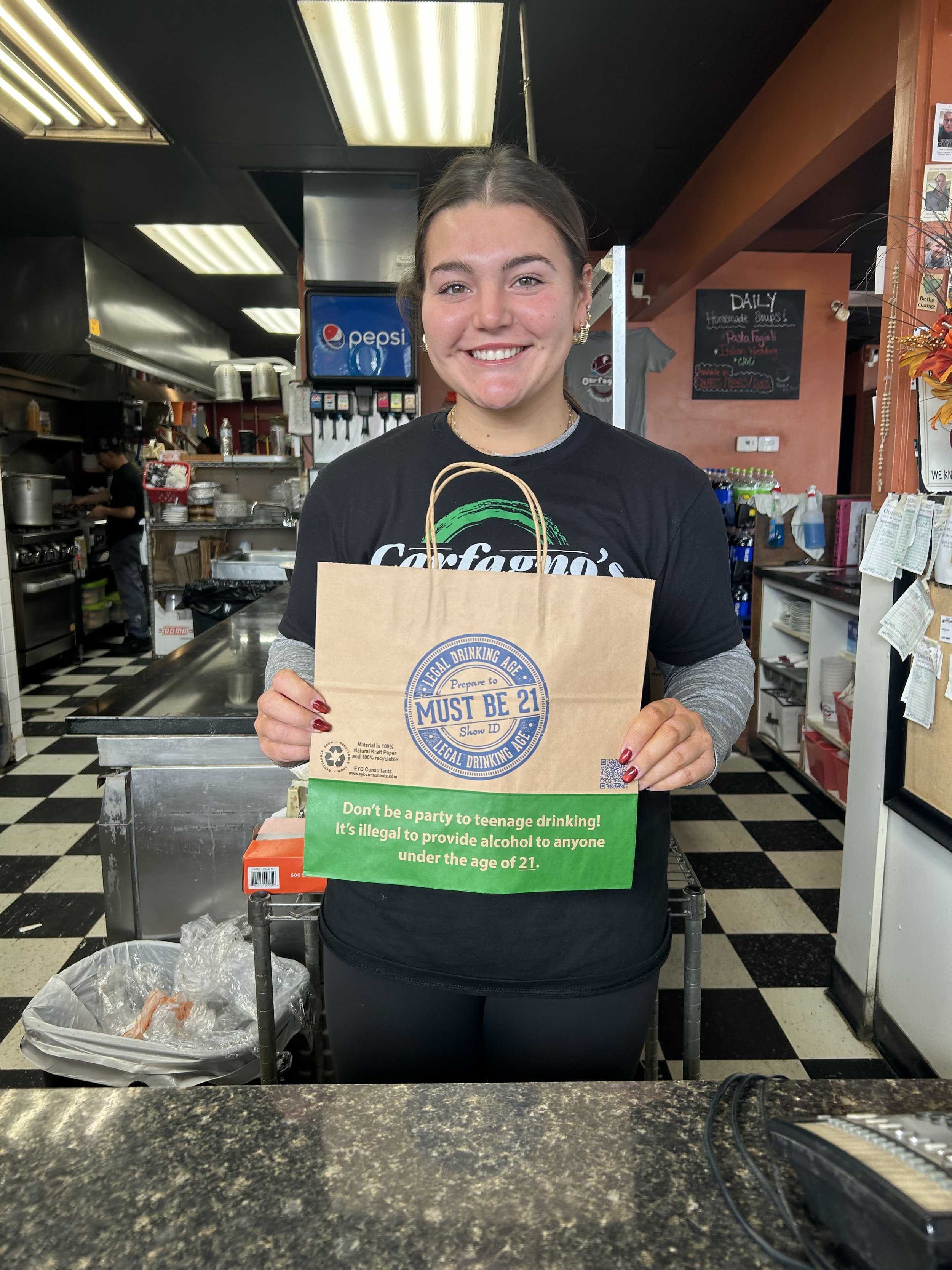 A young woman smiling and holding a paper bag with a age restriction label in a restaurant or cafe setting.