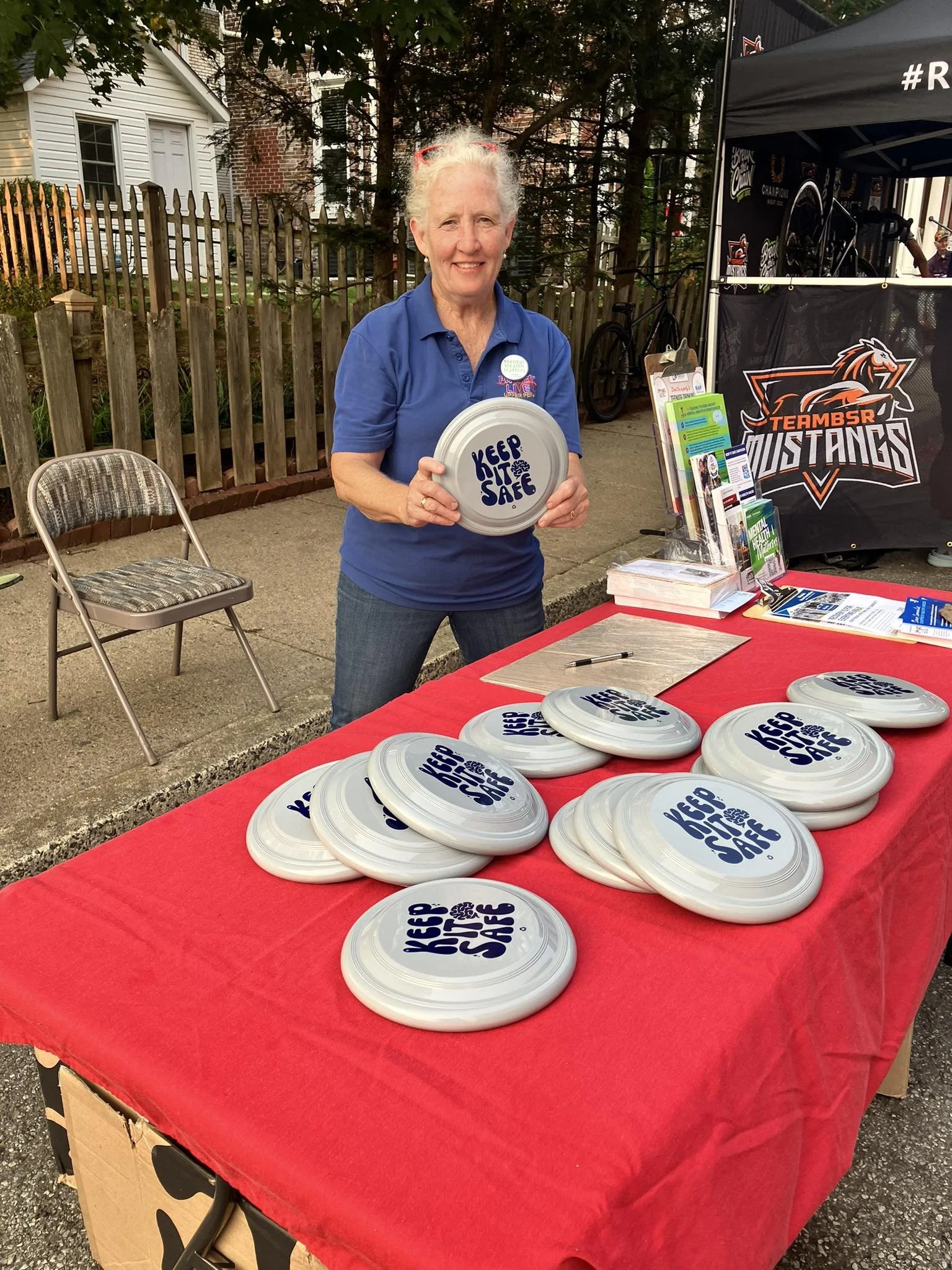 A woman standing behind a table with multiple white frisbees that say "Keep it Safe" on a red tablecloth. She is holding one frisbee and smiling at the camera. The setting appears to be outdoors, with a fence, trees, and a tent with "Mustangs" brandi
