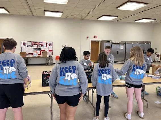 Group of children in a cafeteria or school room, wearing matching gray sweatshirts with the slogan 'Keep It A Secret' printed on the back, standing around a table with food and drinks.