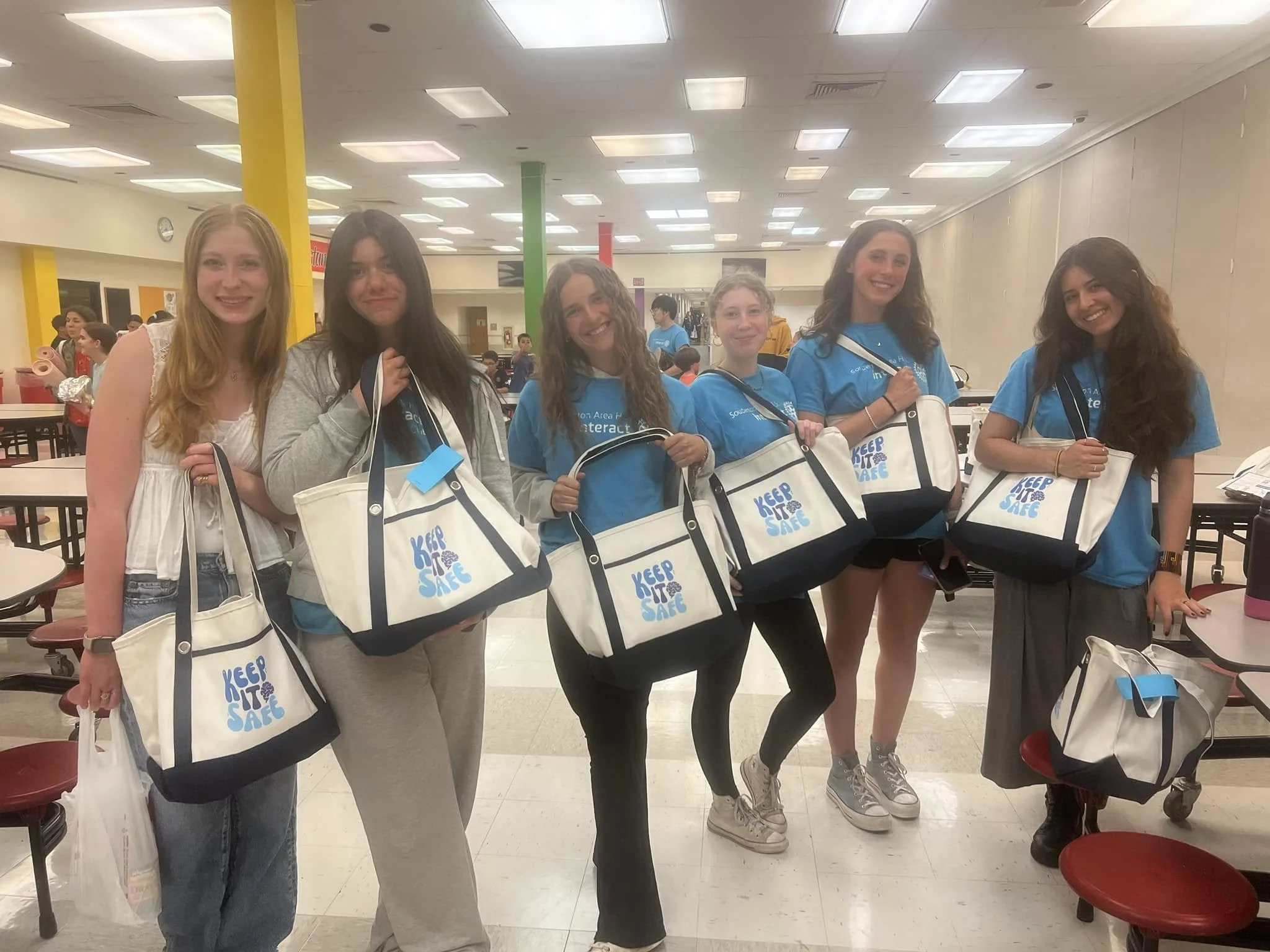 Six girls standing in a cafeteria, holding matching tote bags that say 'Keep It Safe,' smiling at the camera. Four girls wear blue shirts, two wear casual clothing.
