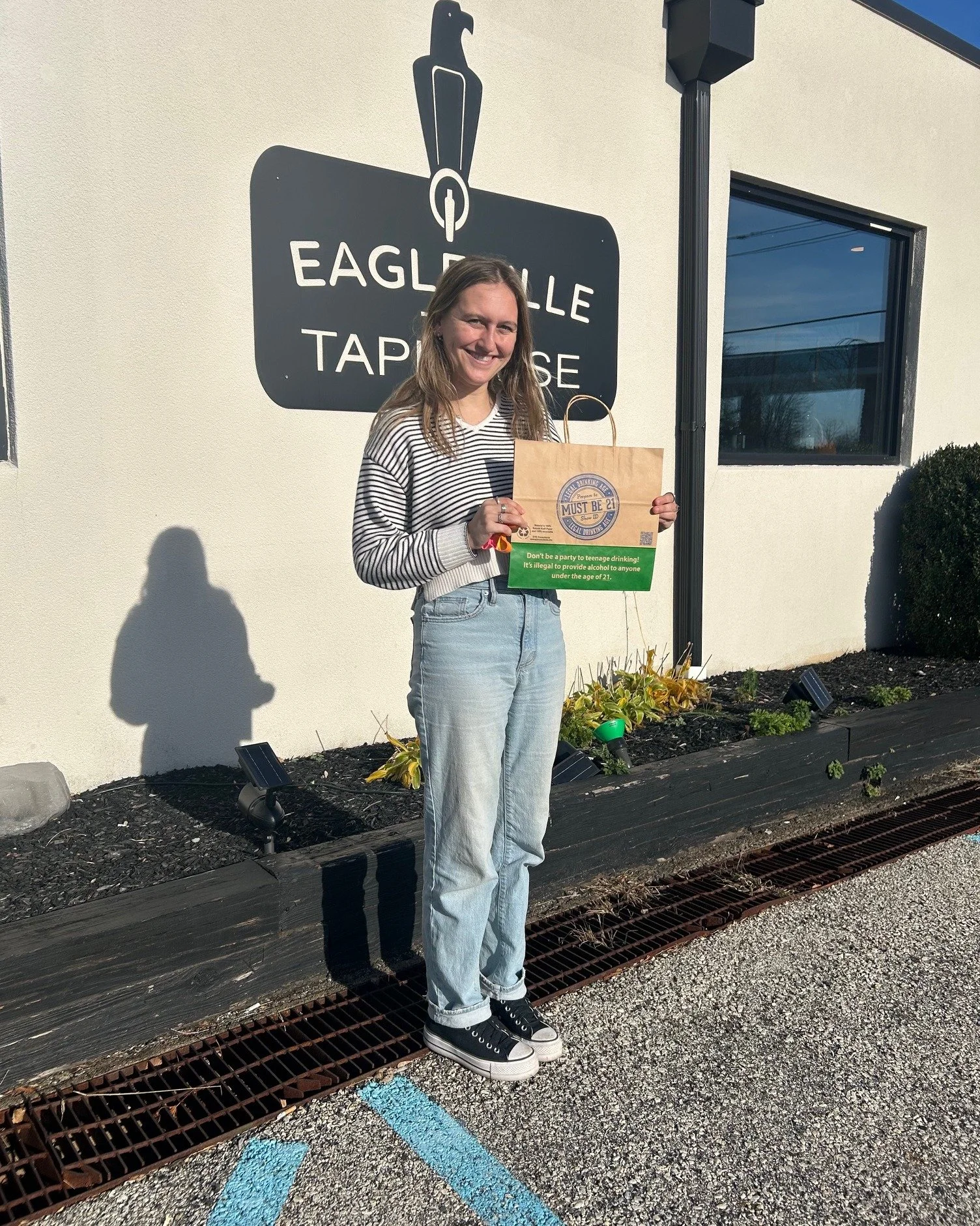 A young woman standing outside Eagleville Tap House, holding a brown paper bag and smiling. She is dressed in a striped sweater, light blue jeans, and black sneakers. The background shows a sign for Eagleville Tap House and a small garden bed with so