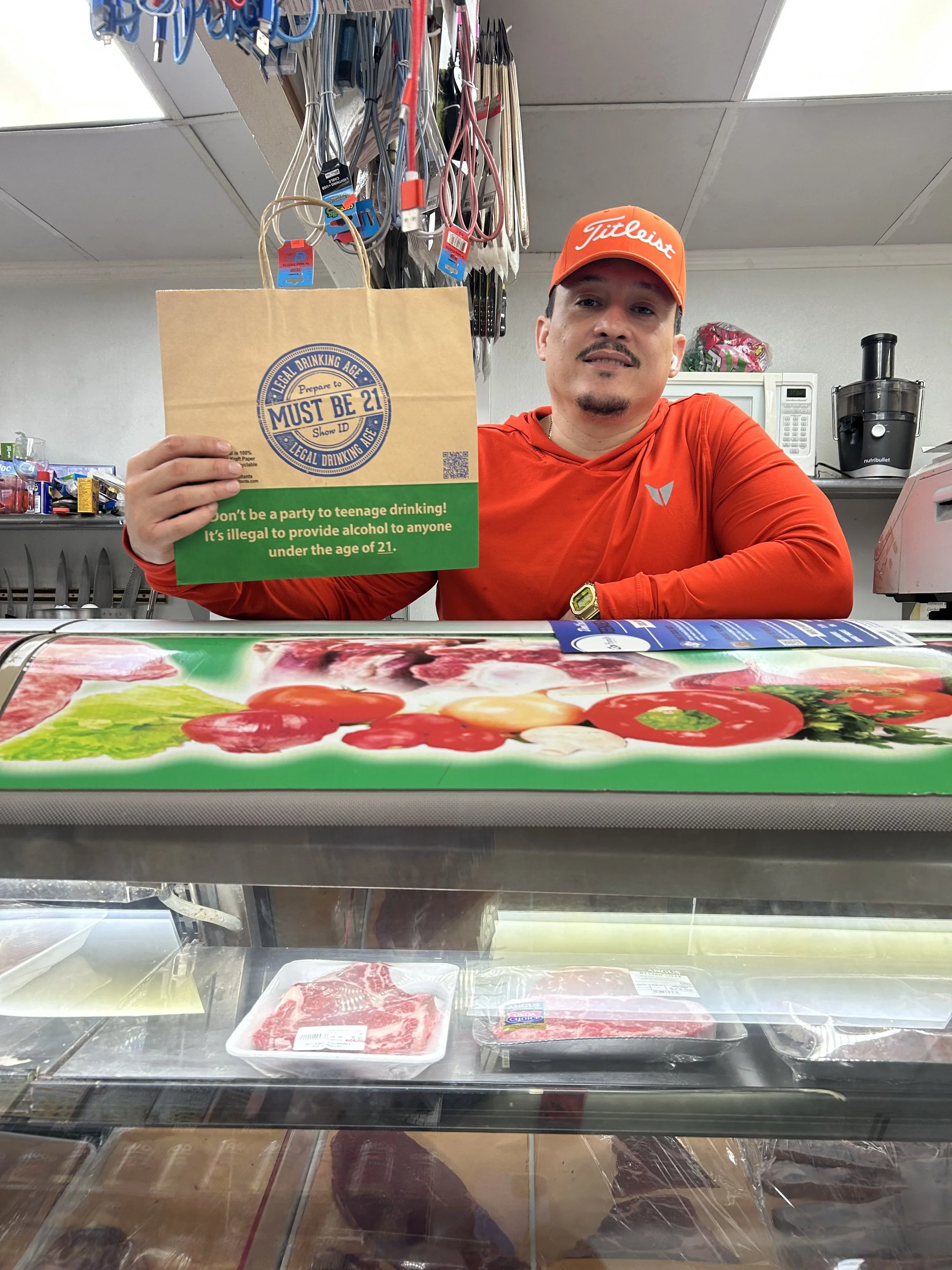 A man wearing a bright red shirt and orange Titleist cap sitting behind a supermarket meat counter. He is holding a paper bag with a green section that has a legal drinking age warning. The counter has packages of raw meat, and there are kitchen uten