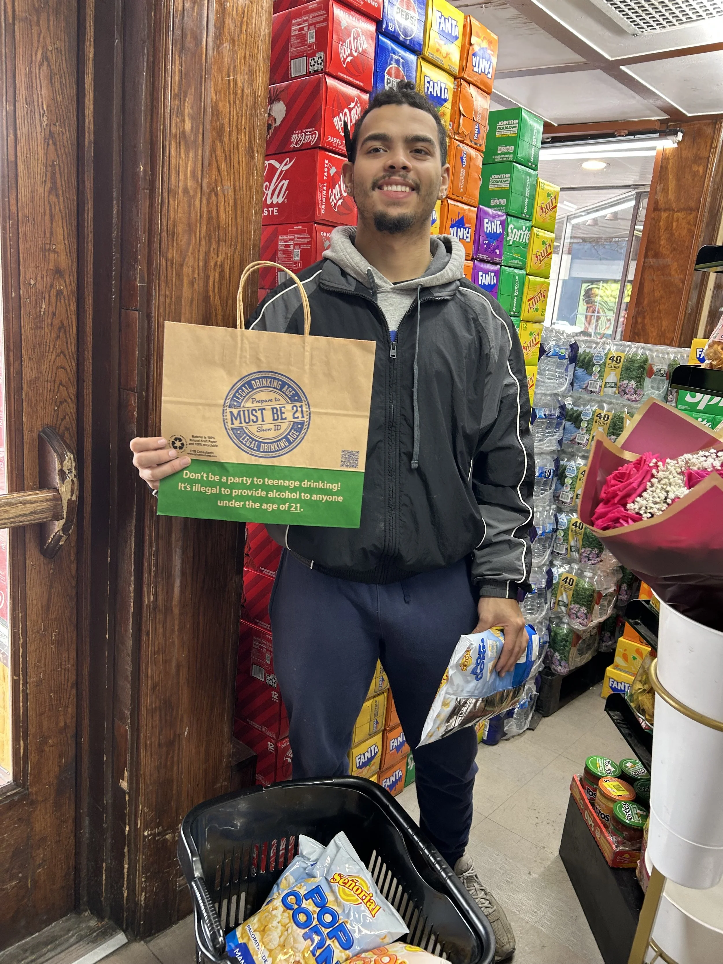 A young man standing in a store aisle holding a paper bag with a 'Must Be 21' sign and a bag of popcorn, next to a shopping cart filled with more bags of popcorn. He is smiling and wearing a black jacket with a gray hoodie underneath, with colorful s