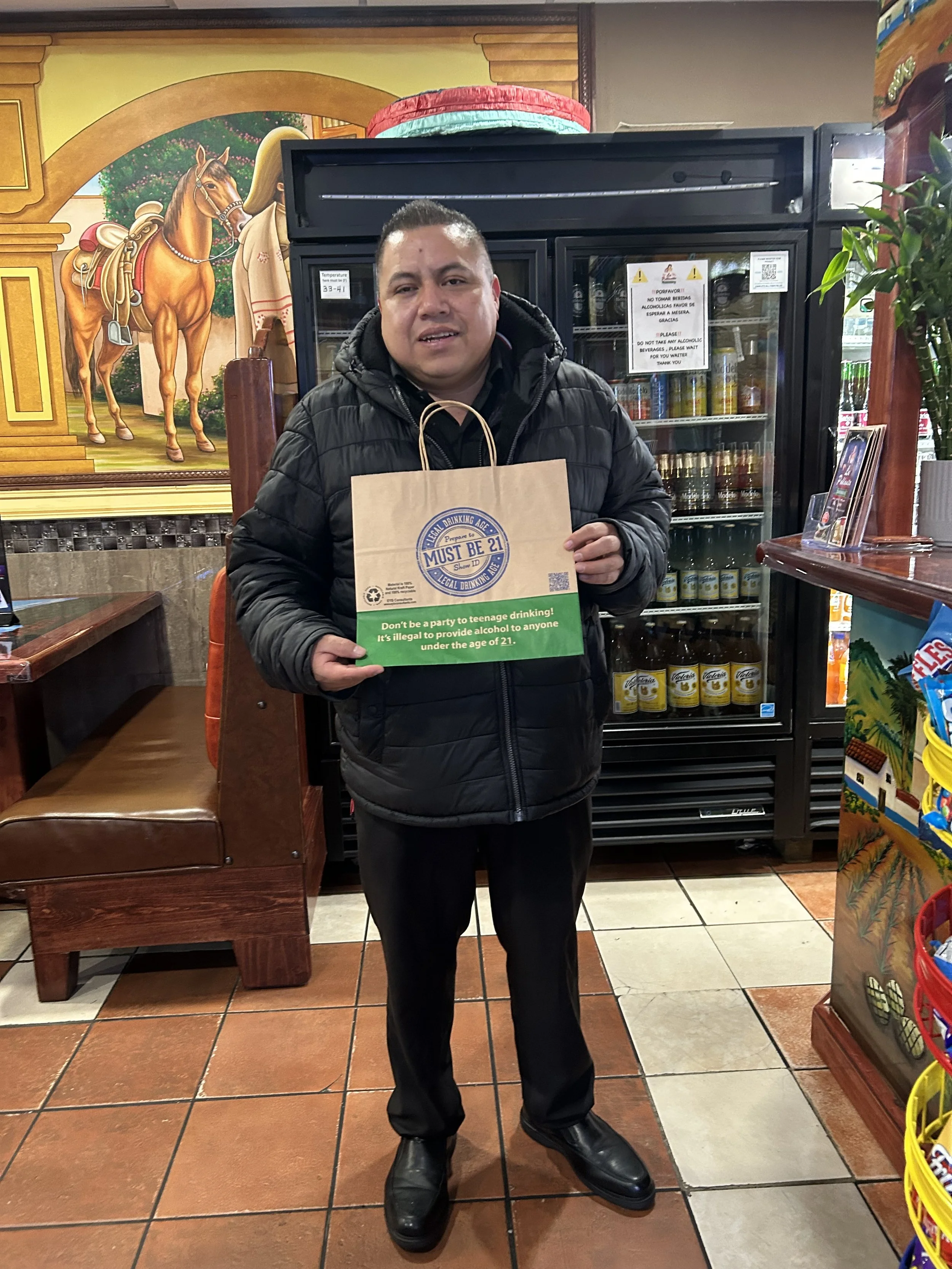 A man standing inside a store holding a paper bag with a green and blue logo. Behind him is a glass-front refrigerator and a colorful mural of a horse and a cowboy. The man is wearing a black puffer jacket and black pants.