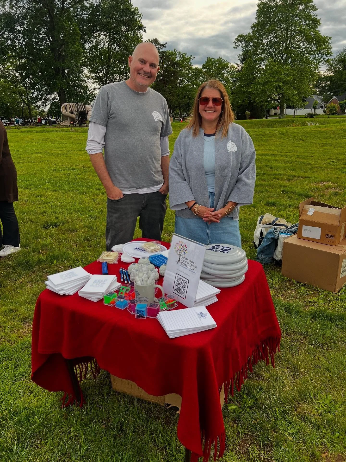 Two people standing behind a table with informational materials at an outdoor event in a park. The table has stacks of notepads, frisbees, and small items. Trees and a playground are visible in the background.