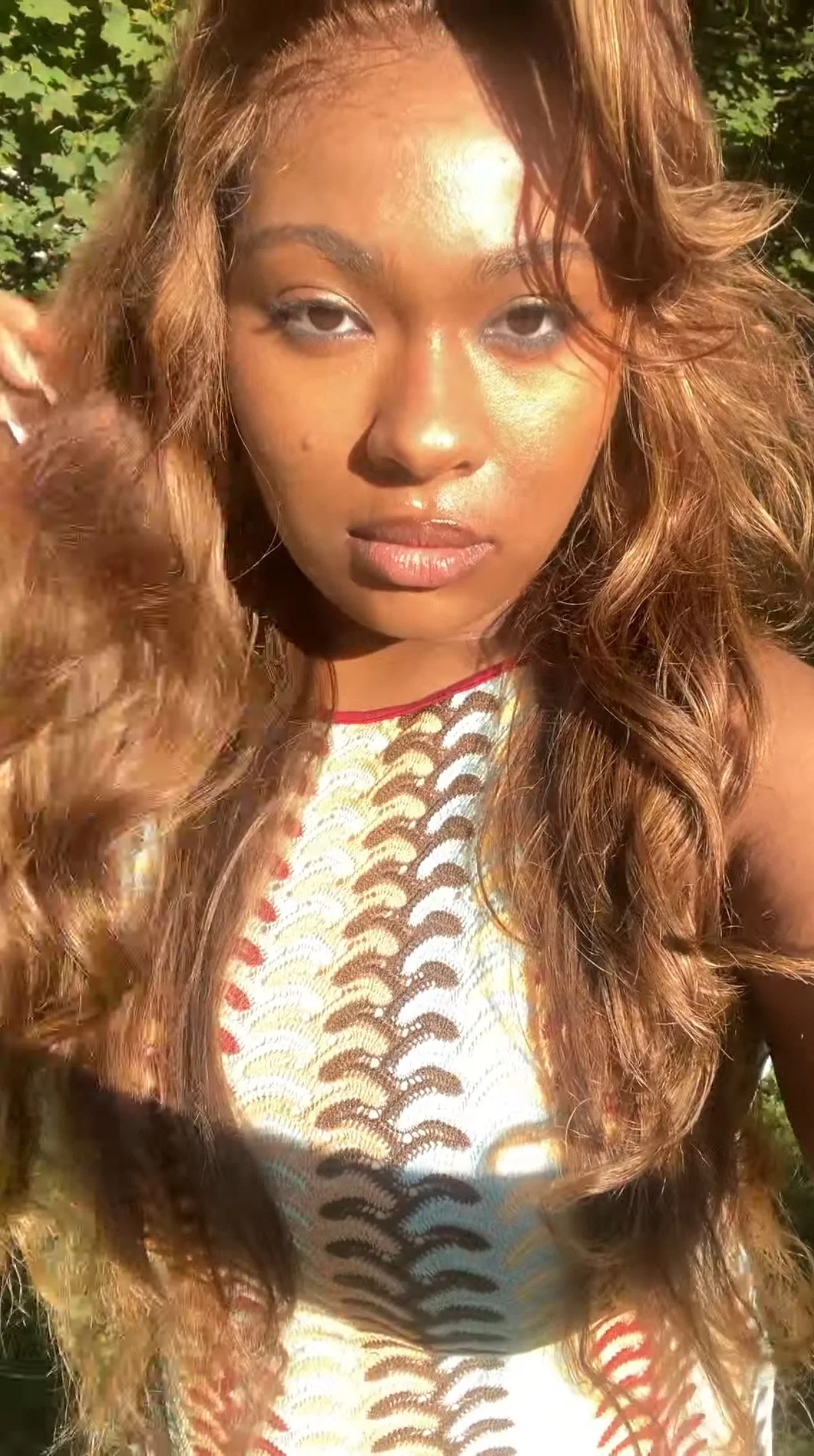 Close-up of a woman with long wavy hair, golden makeup, and a colorful patterned dress, outdoors in sunlight.