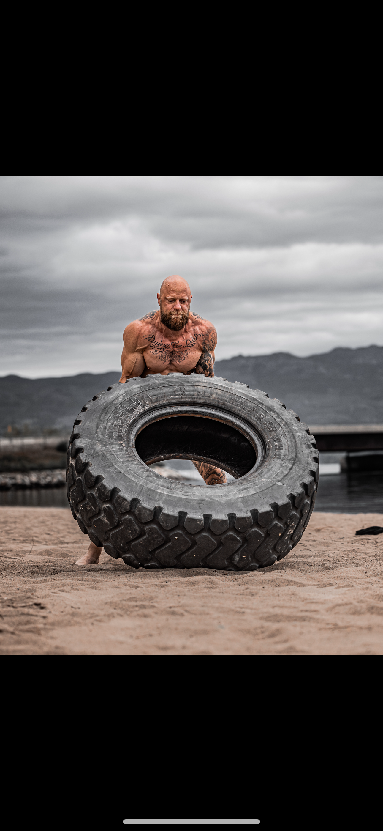 A muscular, tattooed man flipping a large tire on a sandy beach with mountains in the background and cloudy sky overhead.