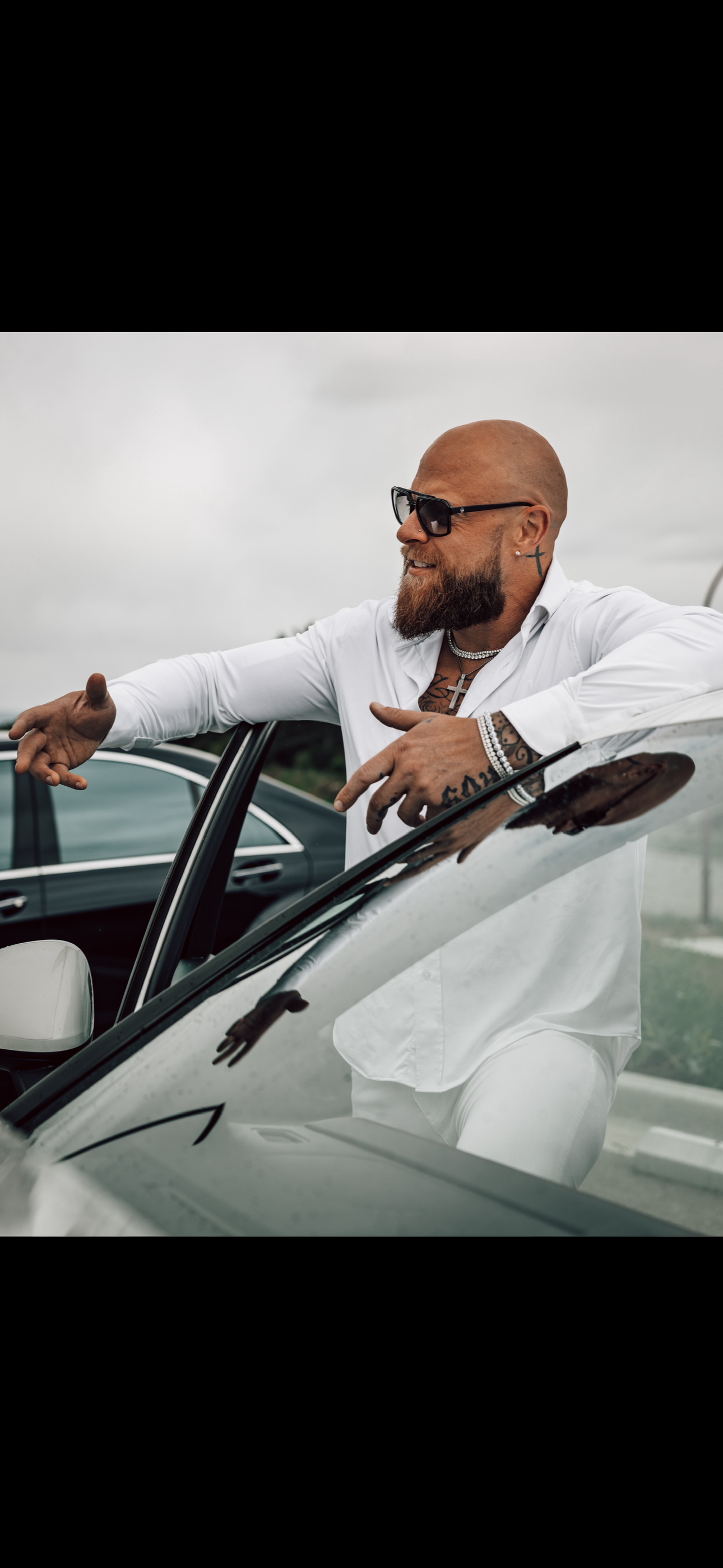 A bald man with a beard, sunglasses, and tattoos, wearing a white shirt and jewelry, leaning out of a white car on a cloudy day.