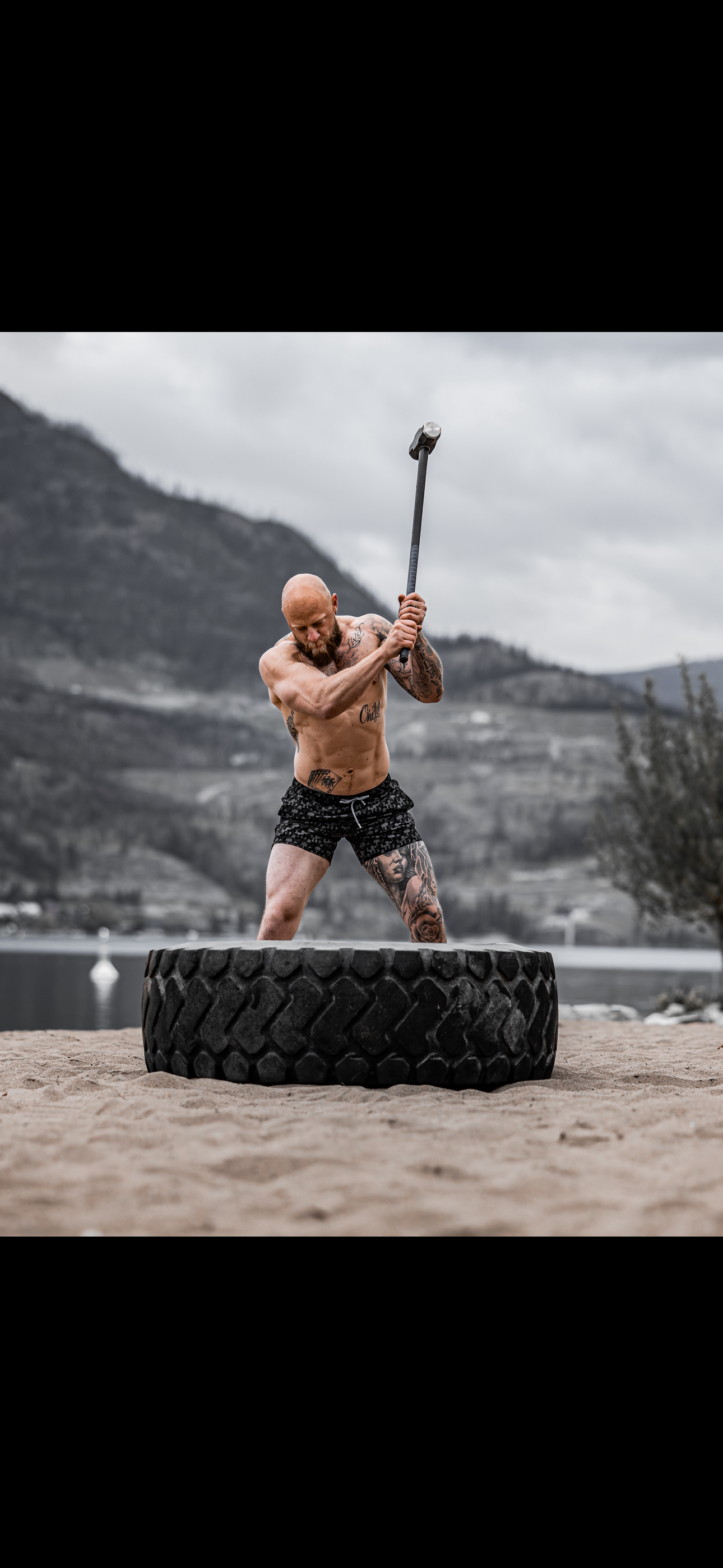 A shirtless tattooed man swinging a sledgehammer onto a large tire outdoors with mountains and a cloudy sky in the background.