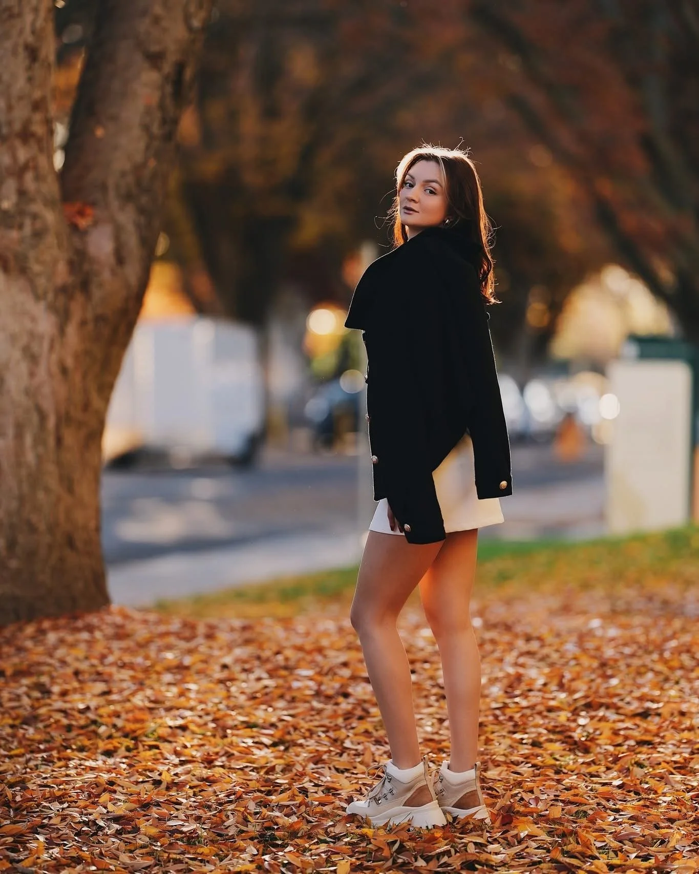 Woman standing on a street covered with autumn leaves, wearing a black coat and white dress.