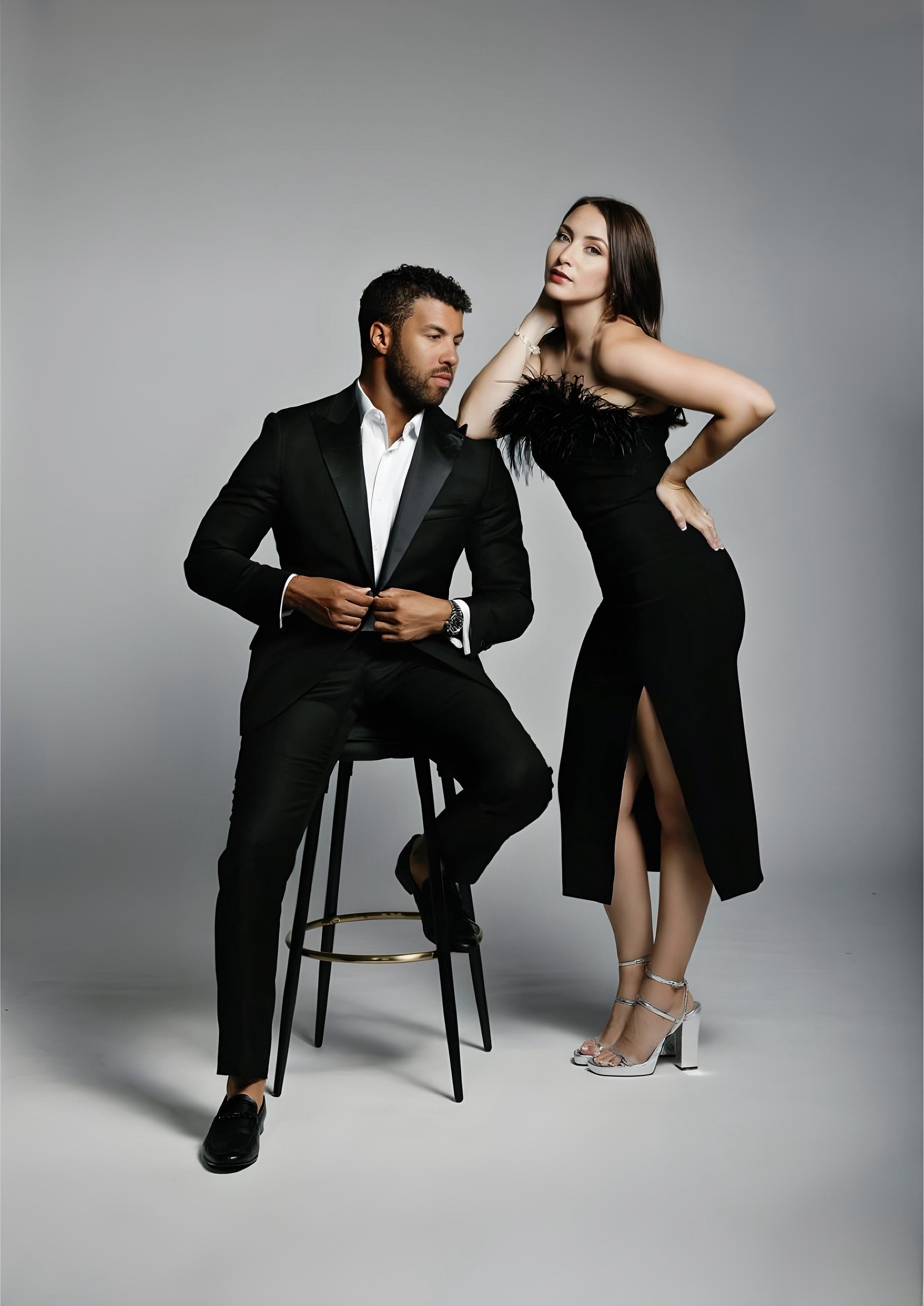 A man in a black suit sitting on a stool next to a woman in a black dress with a feathered top, posing in a studio setting.