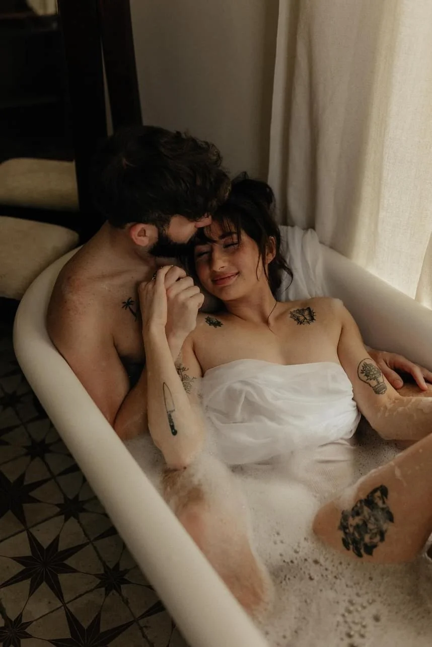 A couple embracing in a bathtub filled with bubbles, with tattoos visible on their arms and shoulders.