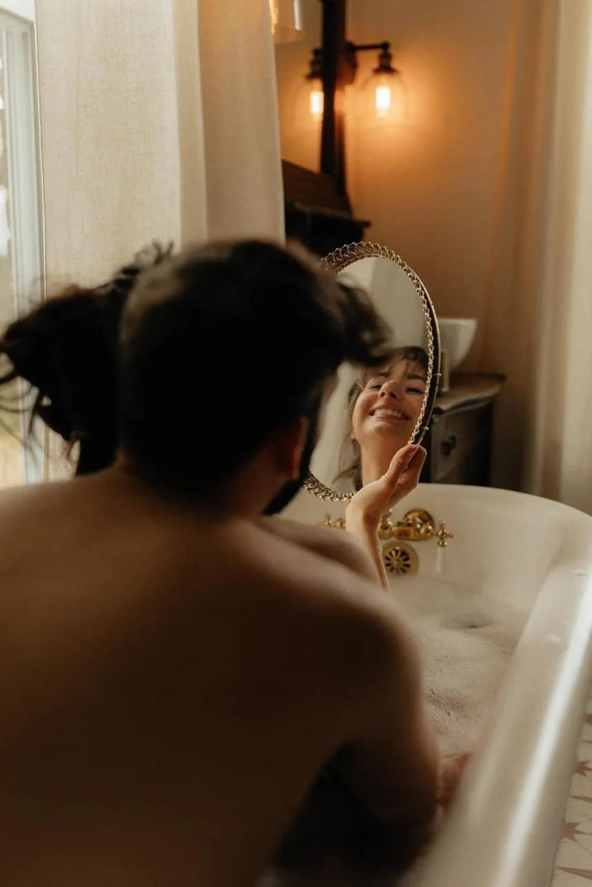 A couple in a vintage bathtub with bubbles, a woman looking into a round mirror, and an illuminated sconce in the background.