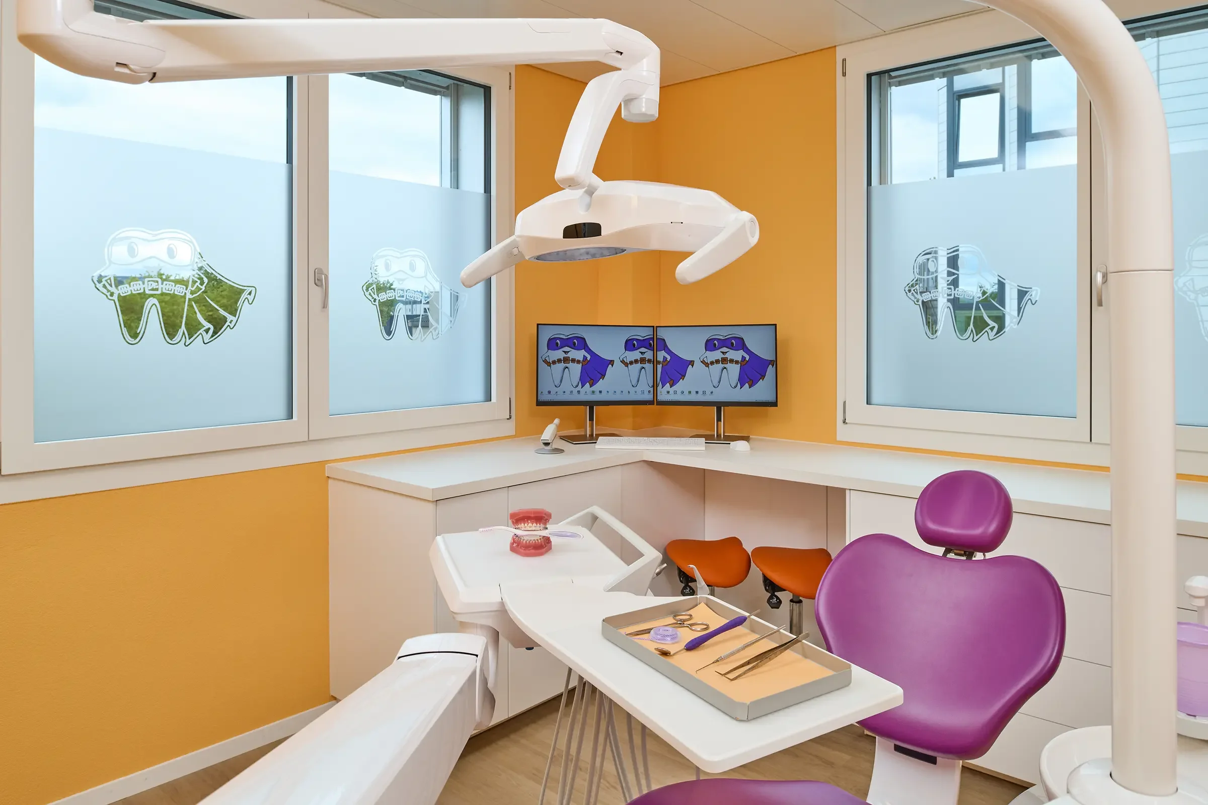 Ein zahnärztliches Behandlungszimmer mit einer Zahnarztliege, einem Behandlungsstuhl mit lila Bezug, einem Behandlungsset mit Werkzeugen, einem Monitor mit Dinosaurier-Grafik, einem Lüfter und Fenstern mit Aufklebern von Dinosauriern.