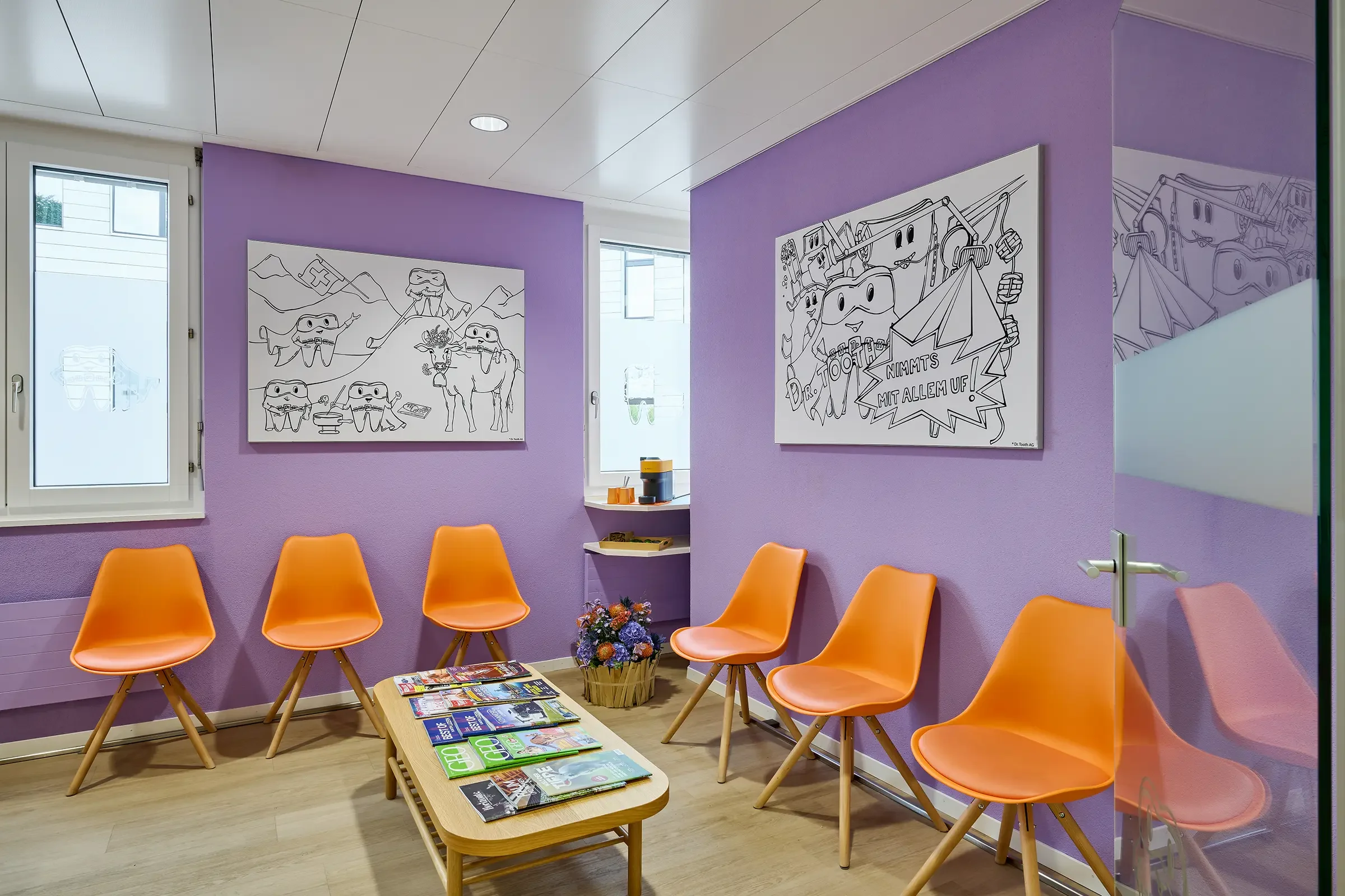 Wartezimmer mit lilafarbenen Wänden, sechs orange Stühlen, einem kleinen Couchtisch mit Magazinen, zwei Fenster, Wandbilder mit Comic- und Cartoon-Darstellungen, Blumenstrauß in Korb, kleines Ablagefach mit Büchern und bunten Gegenständen.