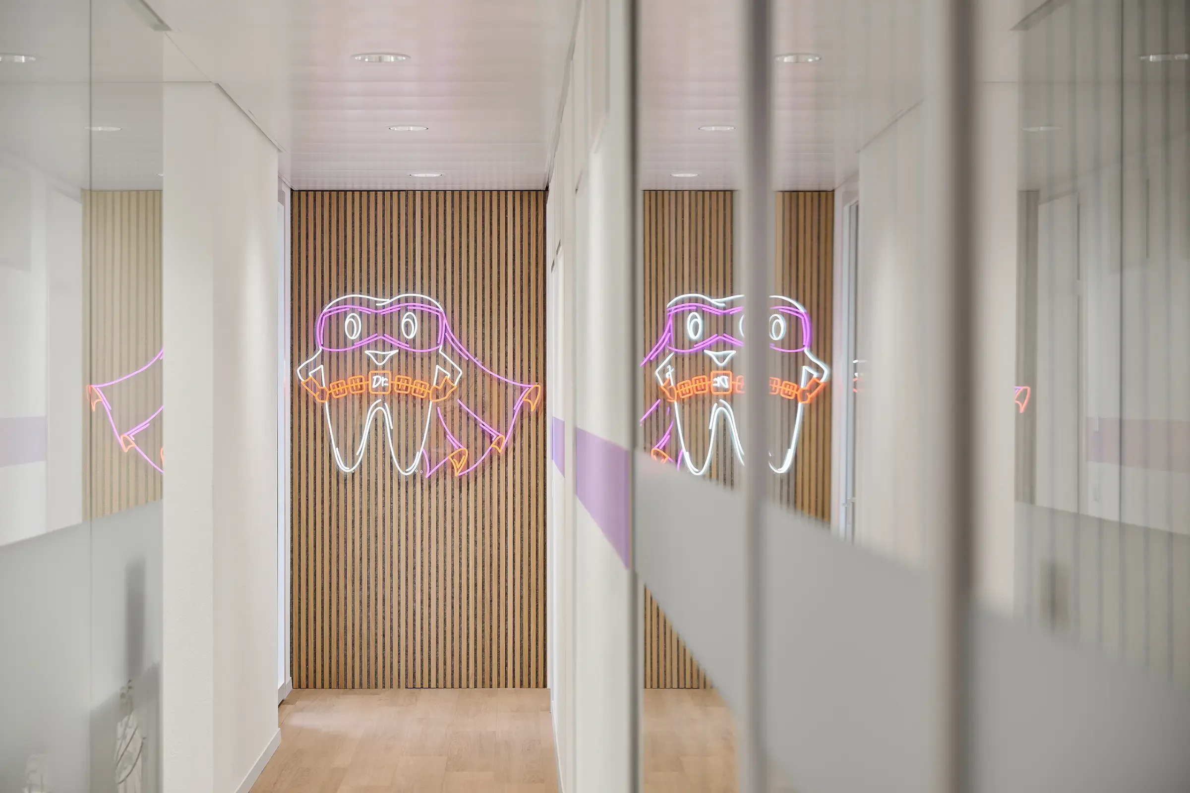 Neon-Licht Illustration eines lachenden Zahns mit Brille und Bisszahnbürste, an einer Holzwaand im Flur.