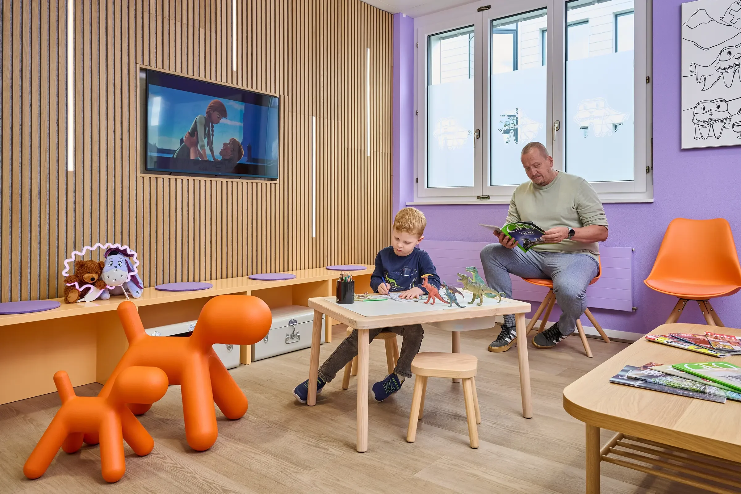 Ein Raum mit bunten Wänden, in dem ein Mann und ein Kind spielen und lesen. Es gibt Spielzeug, einen Fernseher, mehrere Stühle und Zeitschriften.
