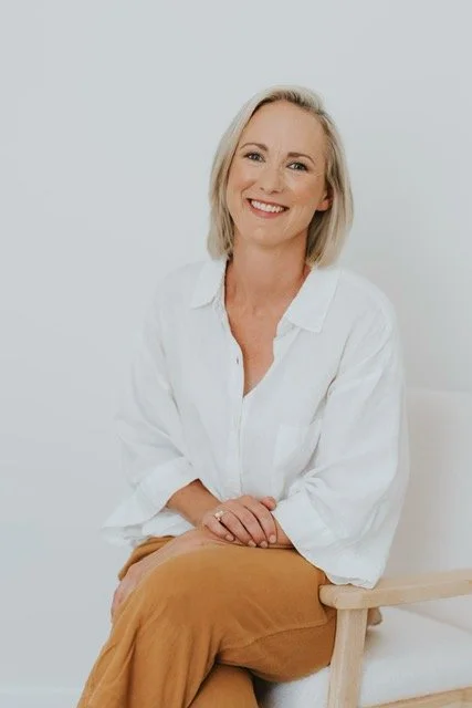 image of Jacinta sitting. She is wearing a white shirt and tan colour pants. Smiling.