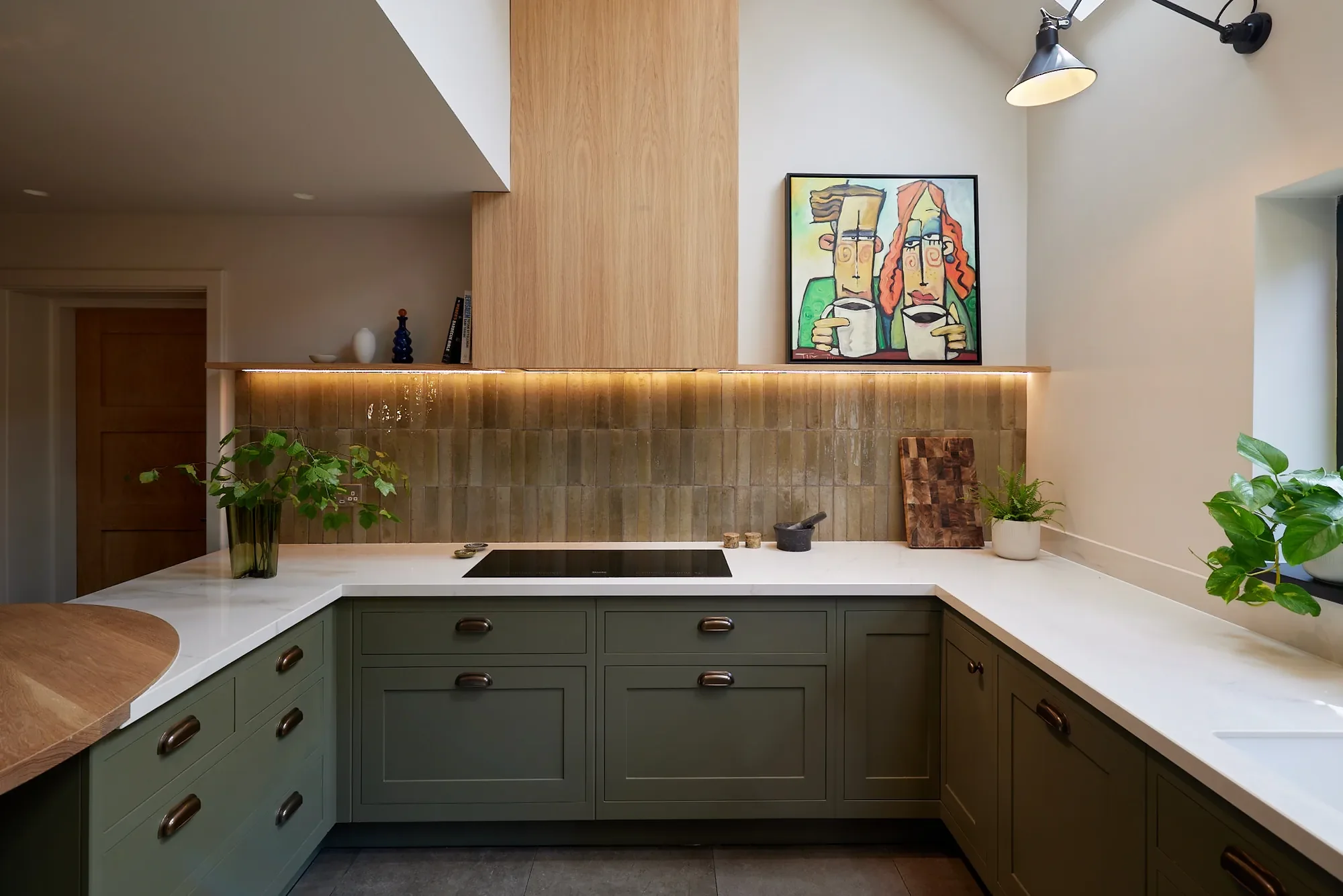 Modern kitchen with green cabinets, white countertop, backsplash, potted plants, and colorful abstract artwork on the wall.