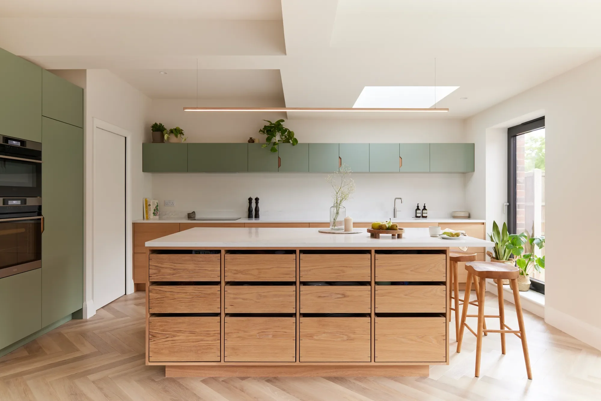 Modern kitchen with green cabinetry, wooden island with storage drawers, white countertop, plants, and natural light from a skylight and window.