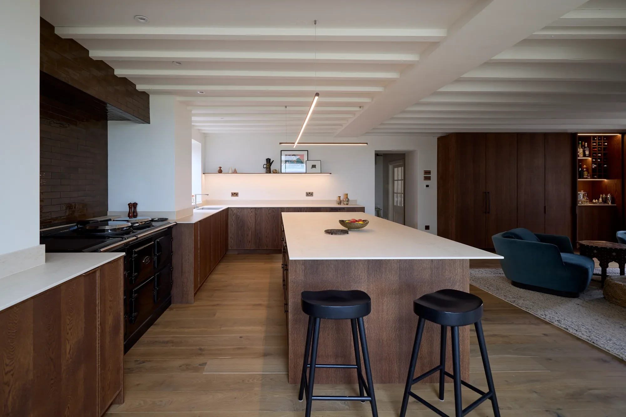 Modern kitchen with a large white island, dark wood cabinets, and a seating area with black stools and a blue armchair. Open layout with wooden floors and minimalist decor.