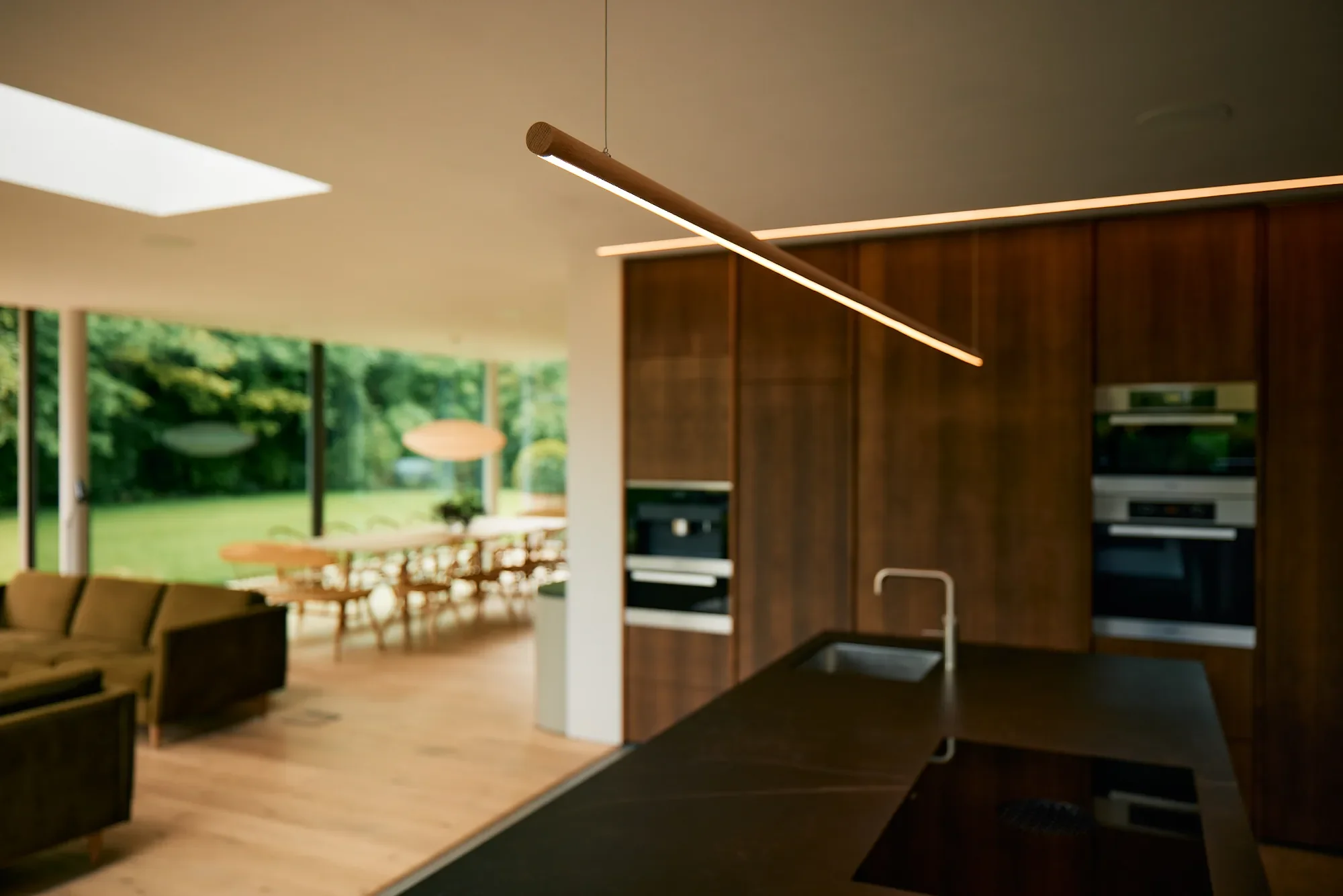 Modern kitchen with dark countertops, wooden cabinets, and built-in appliances, leading to a bright dining and living area with large windows showing a green outdoor space.