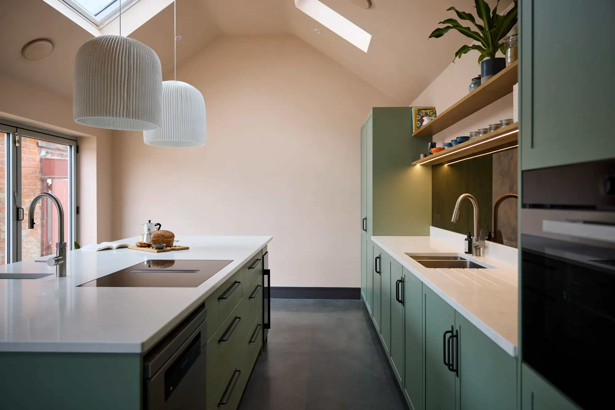 Modern kitchen with white countertops, green cabinets, and a sky window on the ceiling. Two pendant lights hang above the island, which has a loaf of bread, a cup, and an open book. There is a sink with a faucet and a window with a brick wall outside