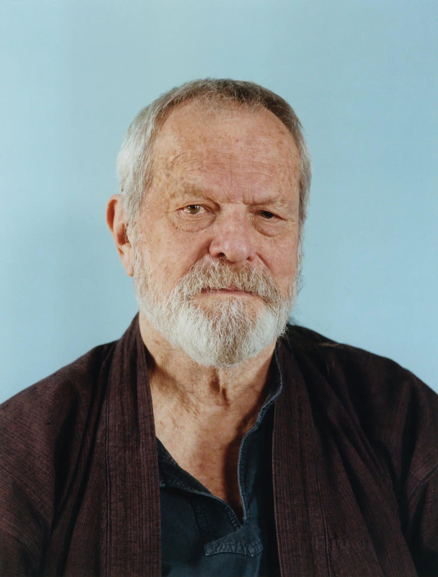 Close-up portrait of Terry Gilliam with gray hair and a beard, wearing a dark shirt and jacket, against a light blue background.