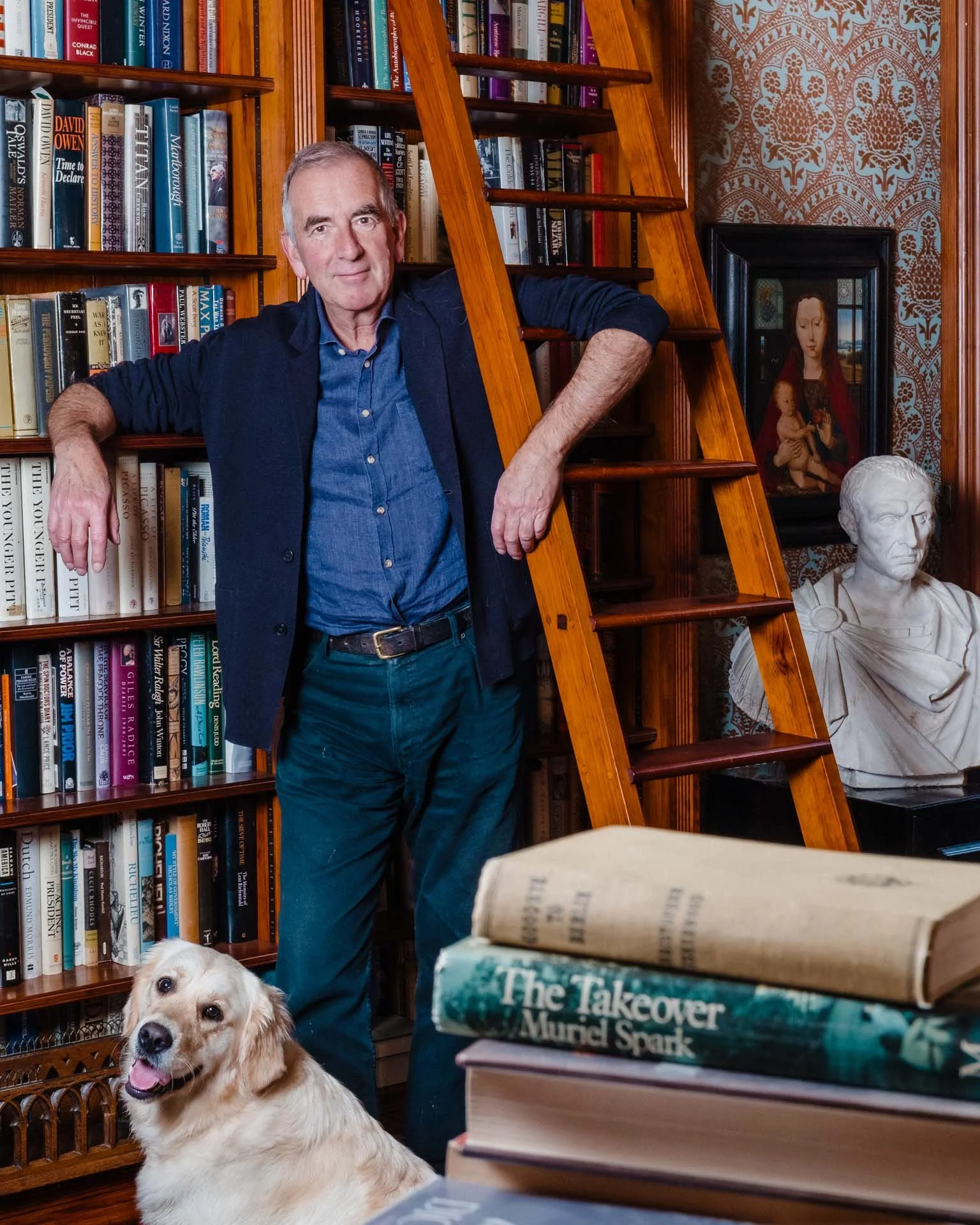 Robert Harris standing next to a wooden ladder in a library, with a golden retriever dog sitting in front of him, and shelves filled with books behind him. There are also framed artworks and a bust sculpture in the background.