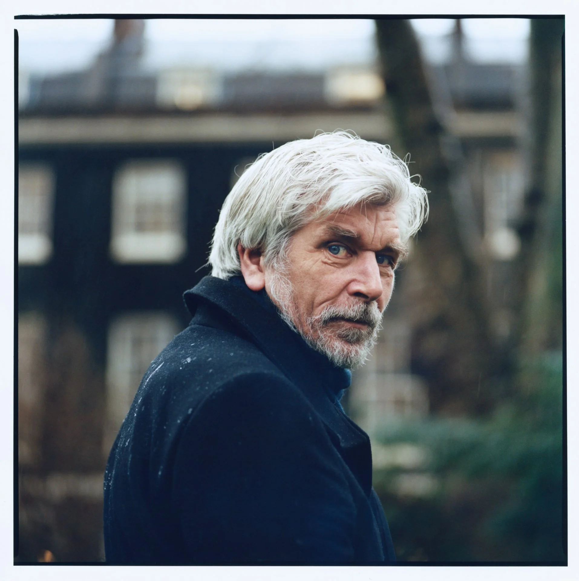 Close-up of Karl Ove Knausgard with white hair, beard, and mustache, wearing a dark coat, looking over his shoulder outdoors with a blurred house and trees in the background.