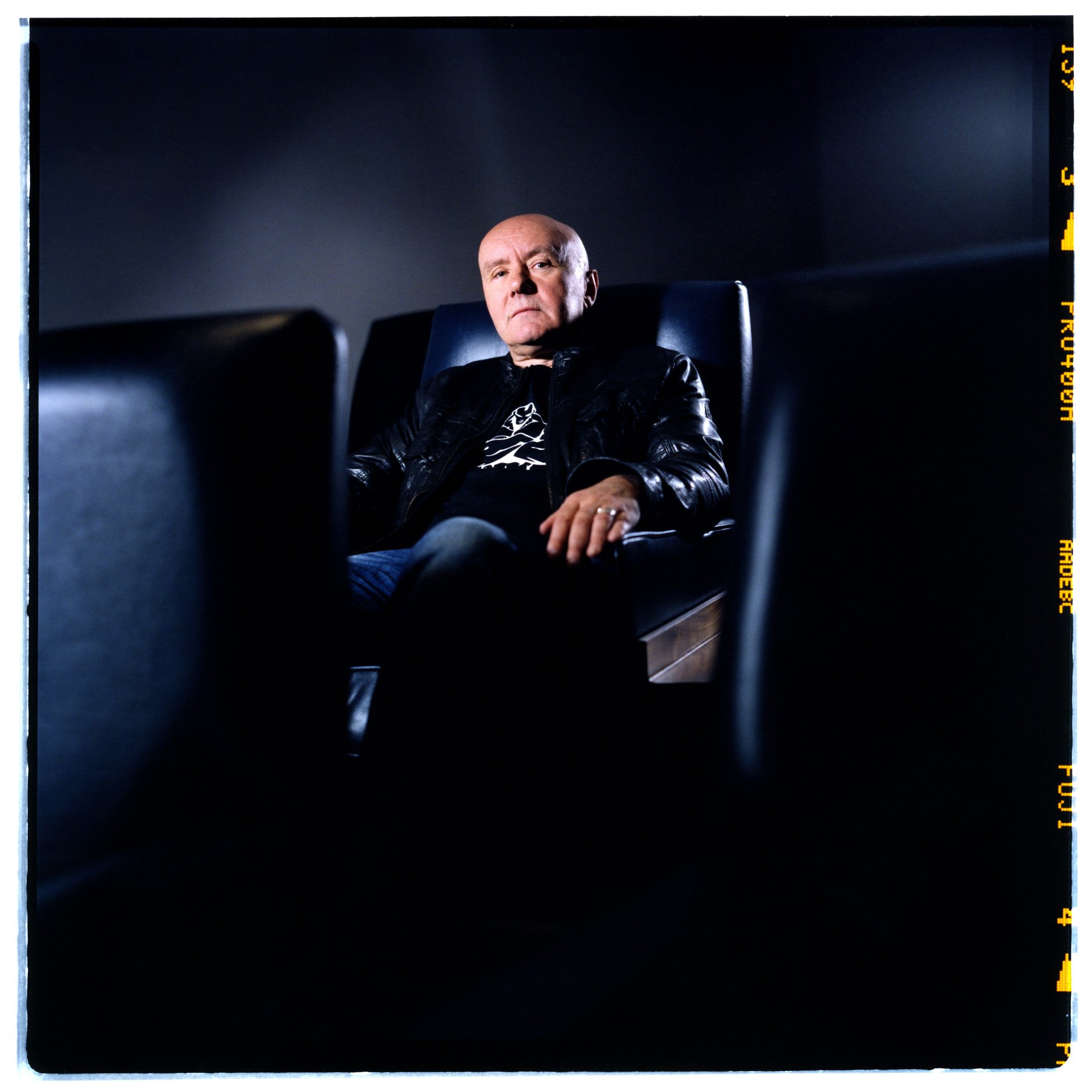 Irvine Welsh wearing a black leather jacket sitting in a dark room on a black leather chair, looking serious, with a dim background and cameras framing the scene.