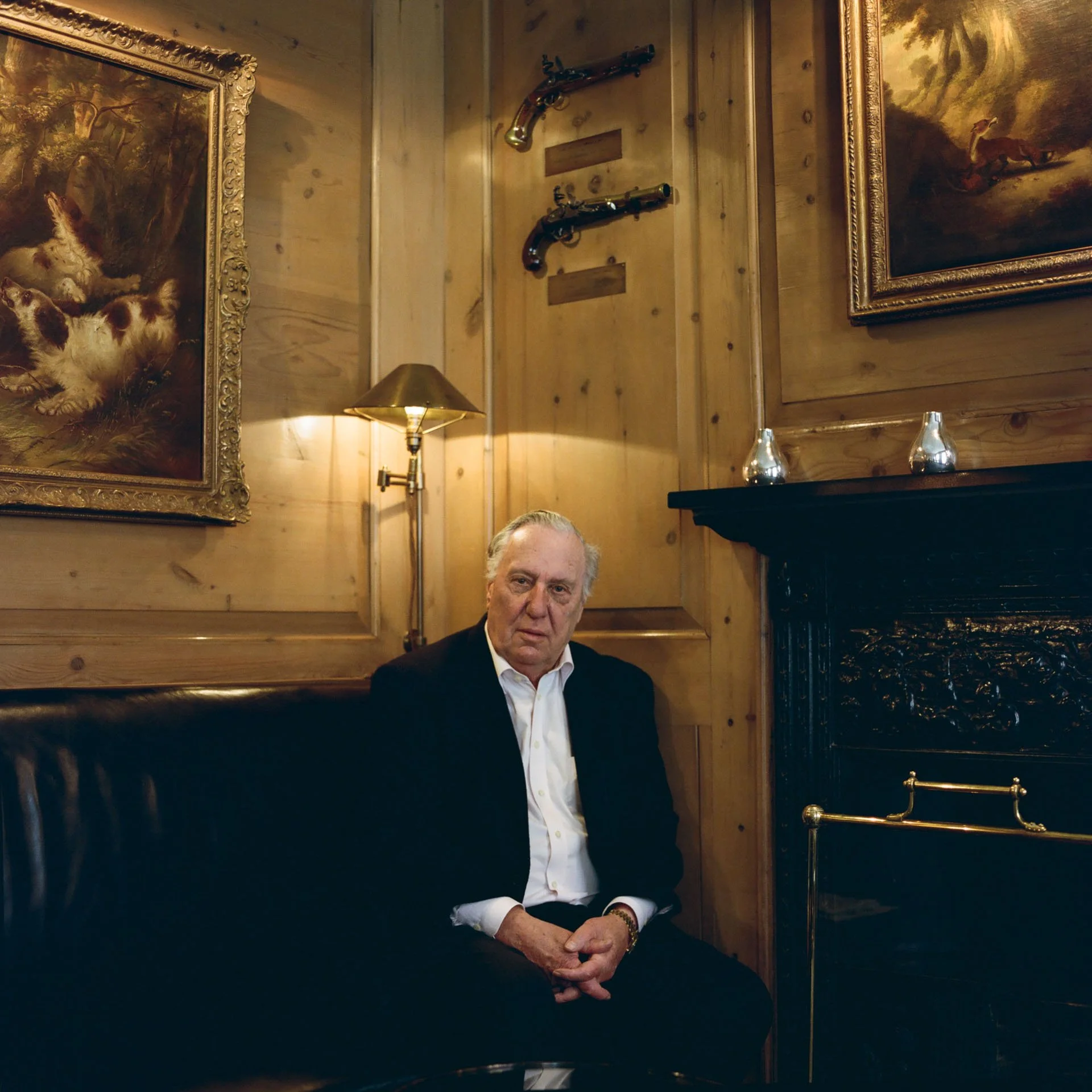 Frederick Forsyth in a black suit and white shirt sits on a black leather bench in a wood-paneled room. The room has paintings, a fireplace, a lamp, and decorative vintage rifles mounted on the wall.