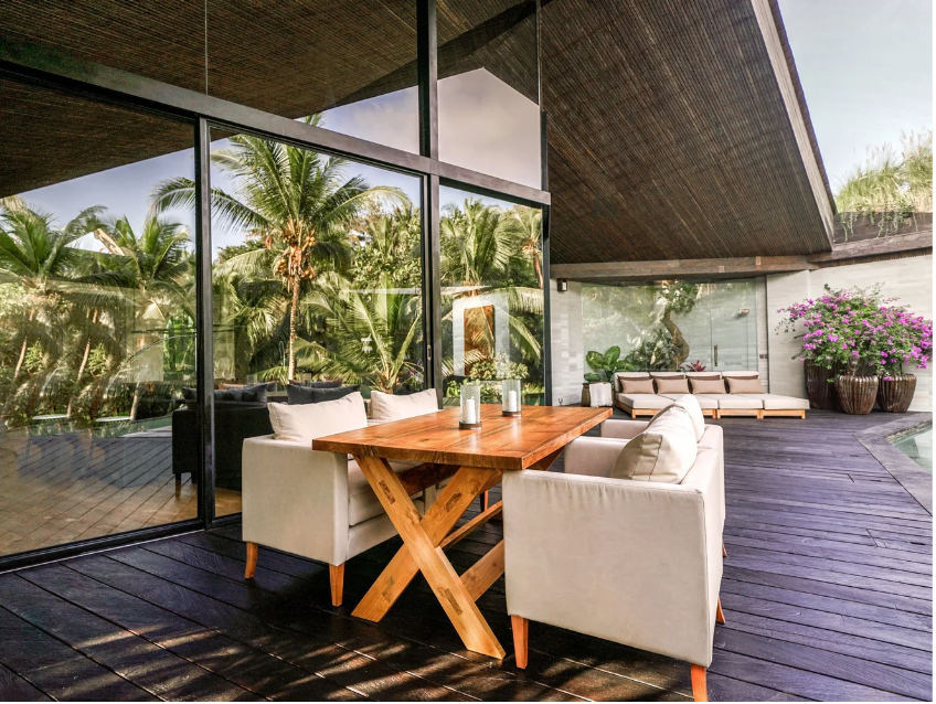Indoor-outdoor living space with large glass windows, wooden deck, white seating, and tropical plants including palm trees and flowering bushes.