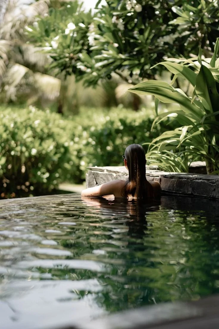 Woman with long brown hair and sunglasses relaxing in a pool surrounded by lush green tropical plants.