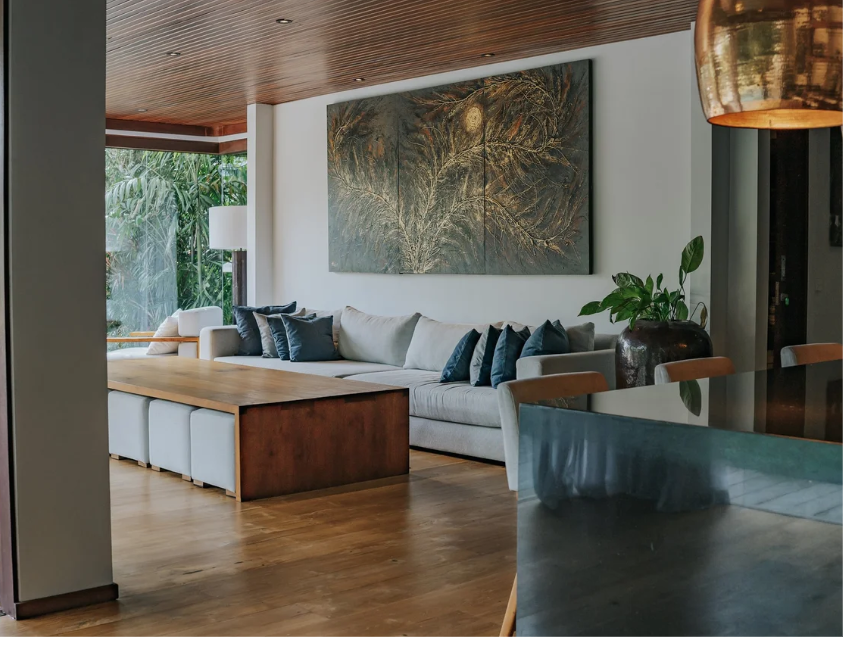 Modern living room with a white sofa adorned with dark blue pillows, a wooden coffee table, large artwork depicting tree branches, a potted plant, and a view of lush greenery outside through a glass window.
