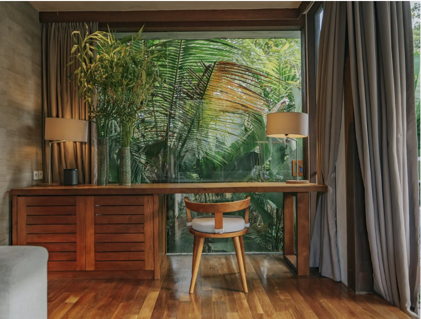 A cozy interior workspace with a wooden desk and a chair, positioned in front of large window overlooking lush green tropical plants outside. The desk has two lamps, one on each side, and built-in storage with wooden doors. The room features hardwood