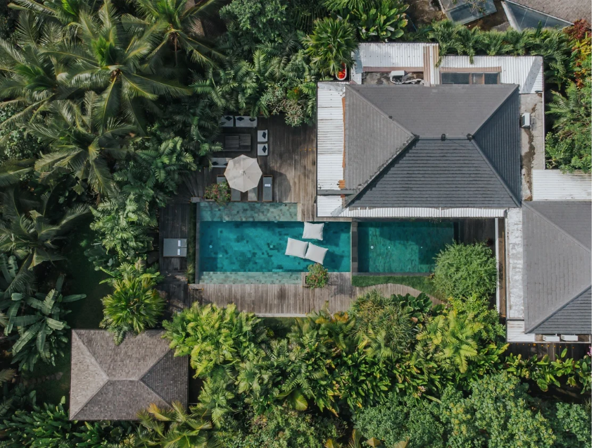 Aerial view of a backyard with a swimming pool, surrounded by tropical plants and trees, with a house and deck area featuring outdoor furniture.
