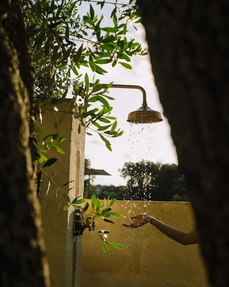 Person reaching out hand to water under an outdoor shower, surrounded by trees and plants.