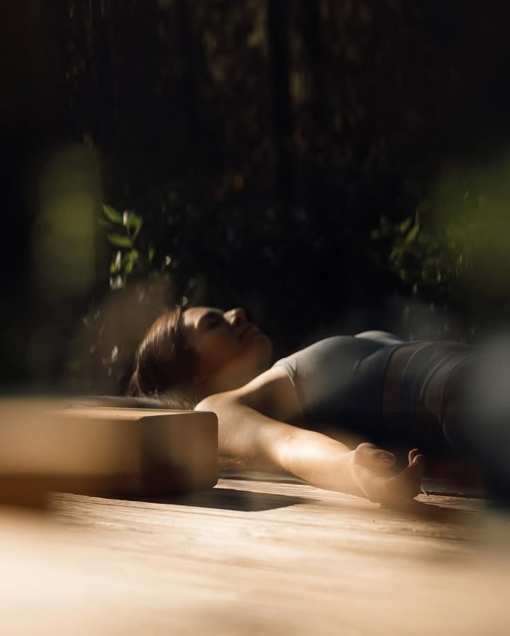 A woman lying on her back on a wooden floor, stretching with her arms extended above her head, in a dimly lit room with plants in the background.