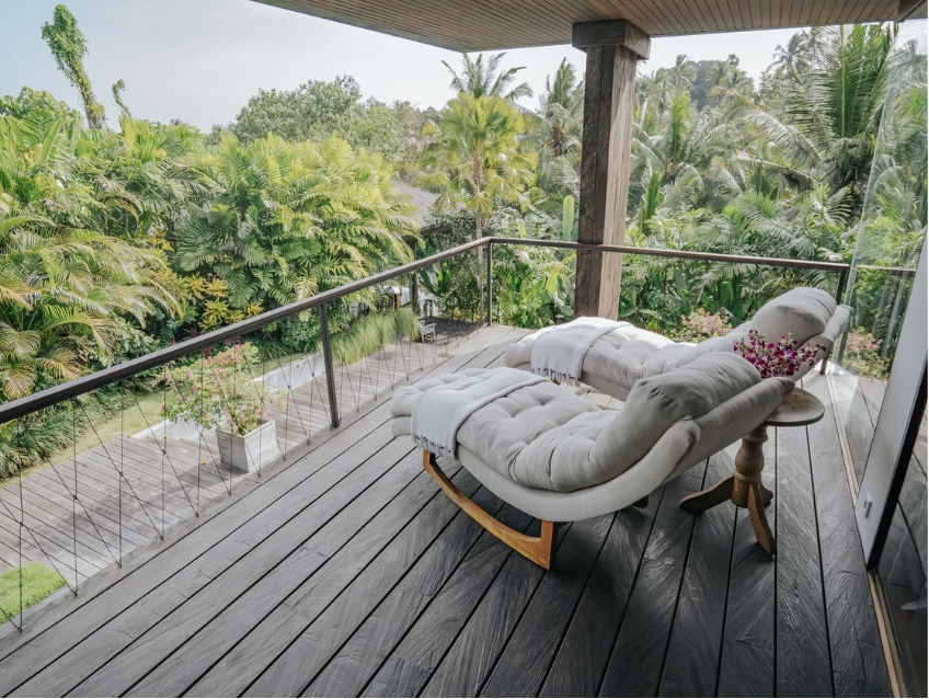 A balcony with wooden flooring and a glass railing overlooking lush green tropical trees and plants. There is a cushioned lounge chair with a white throw blanket and a small wooden side table with flowers on it.