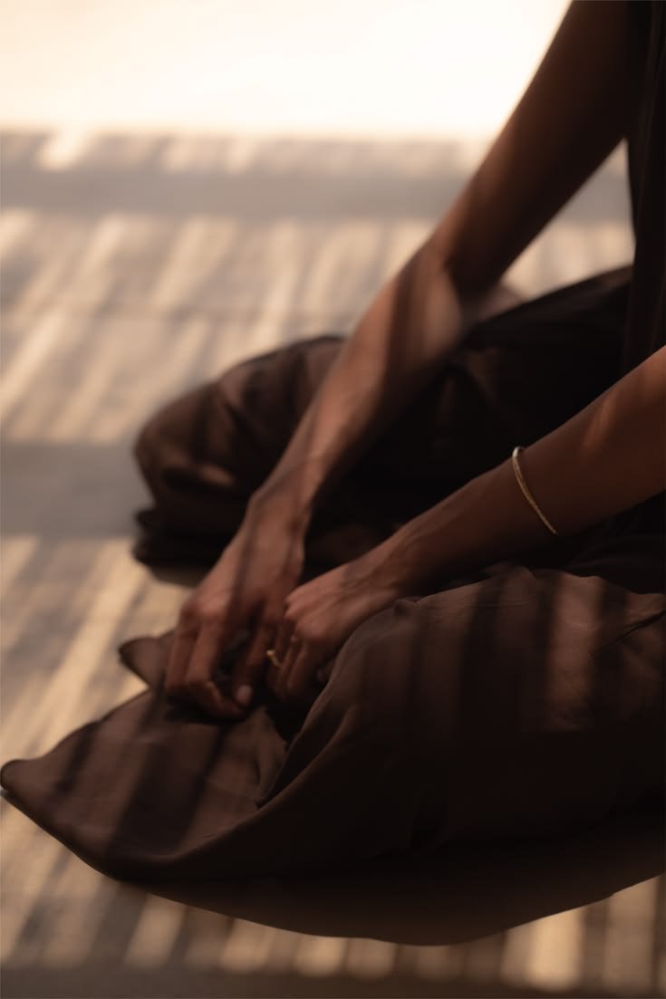 Person sitting on the floor with arms extended, wearing a gold bracelet and a dark outfit, with shadows cast on a wooden floor.