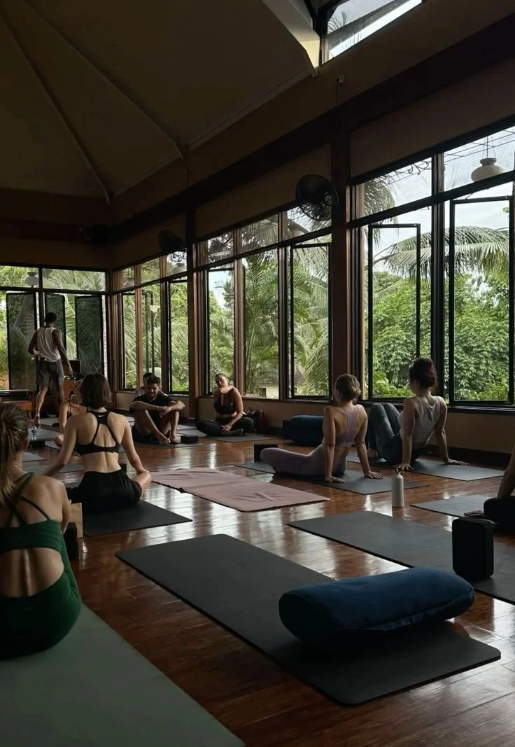 People participating in a yoga class in a wooden-floored studio with large windows showing a lush, green outdoor view.