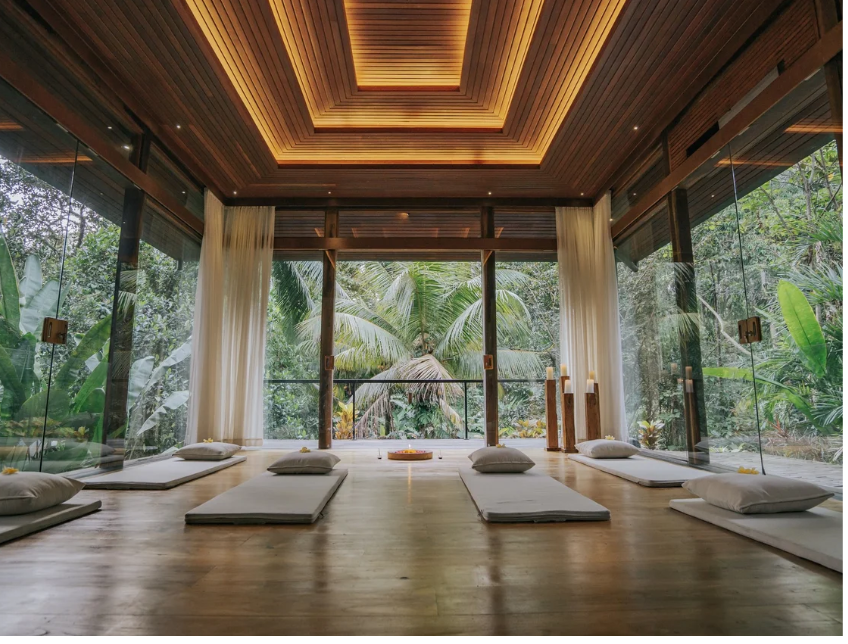 A tranquil meditation space with white cushions and pillows on a wooden floor, surrounded by large glass walls and lush tropical greenery outside.