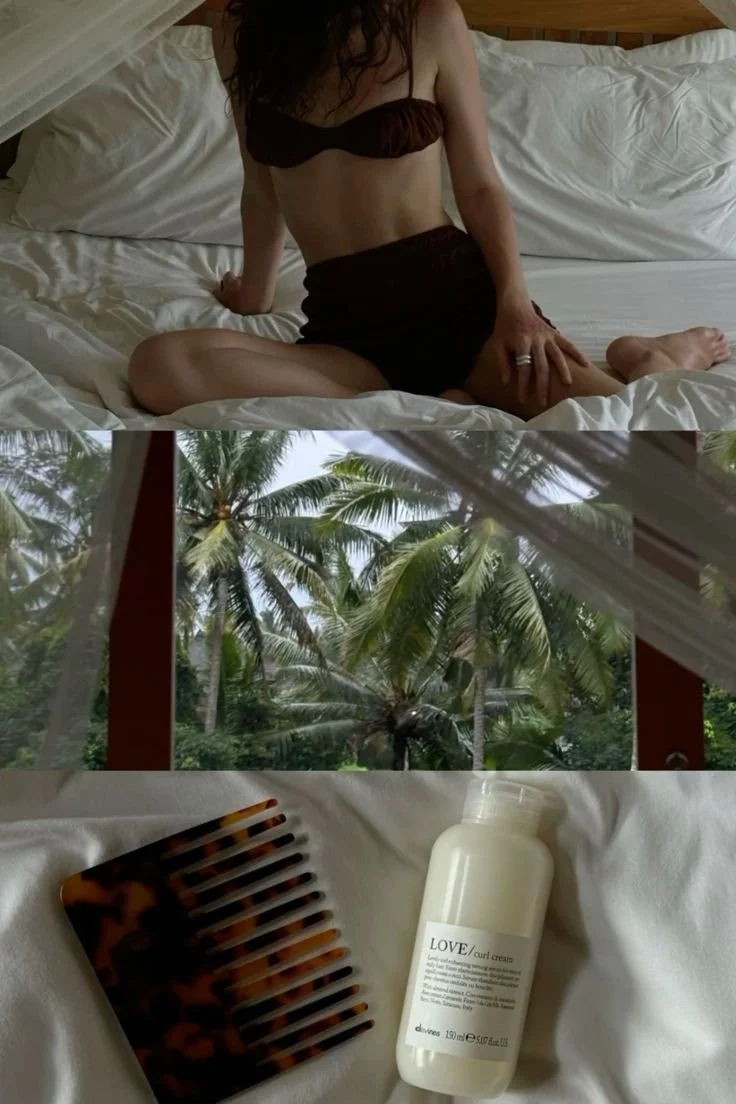 A woman sitting on a bed in a hotel room, a window showing palm trees outside, and a bottle of lotion along with a hair comb on the bed.